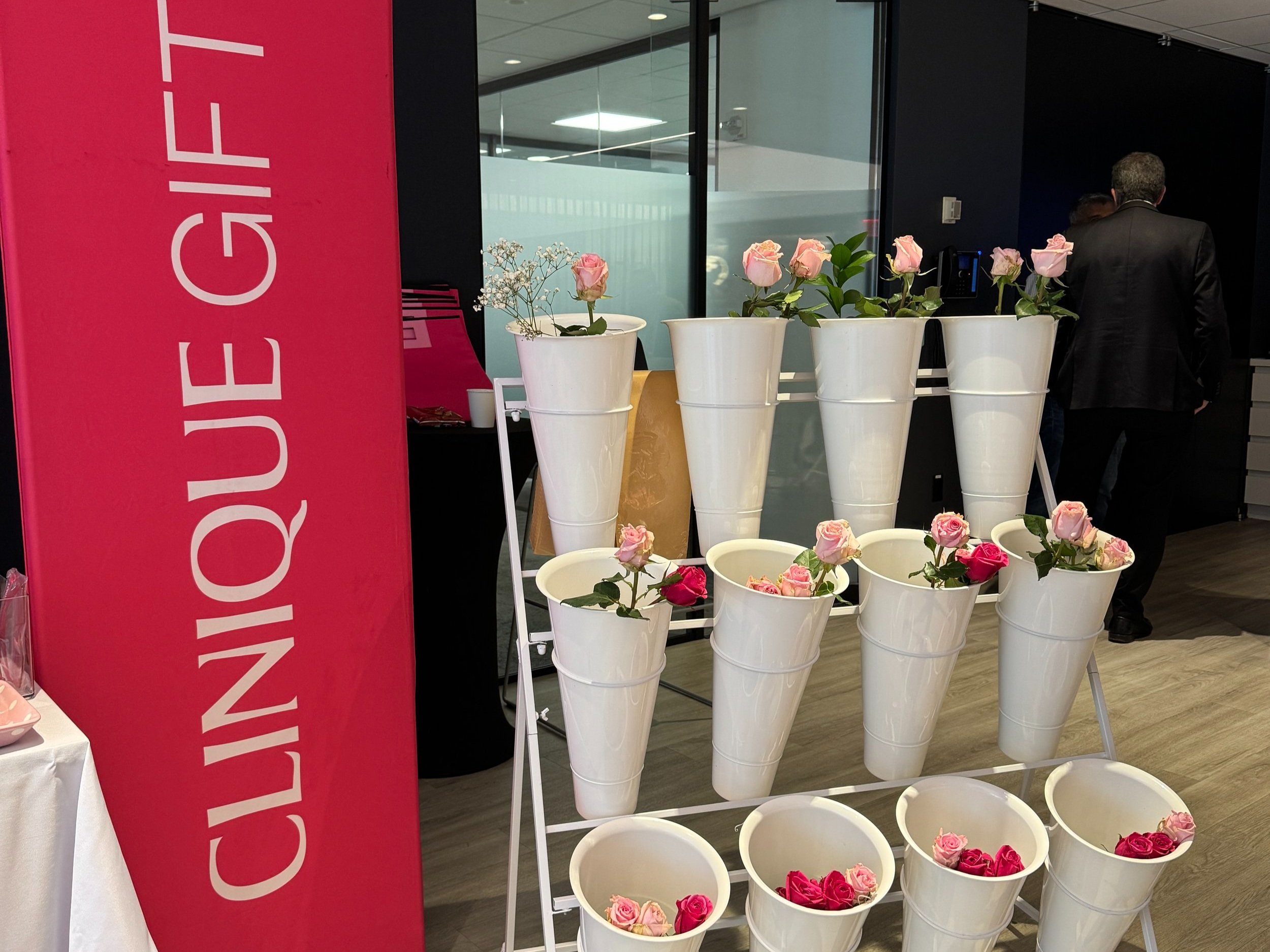 Display of pink and white roses in white buckets, with a red sign that reads ‘CLINIQUE GIFT’ and people in business attire in the background.