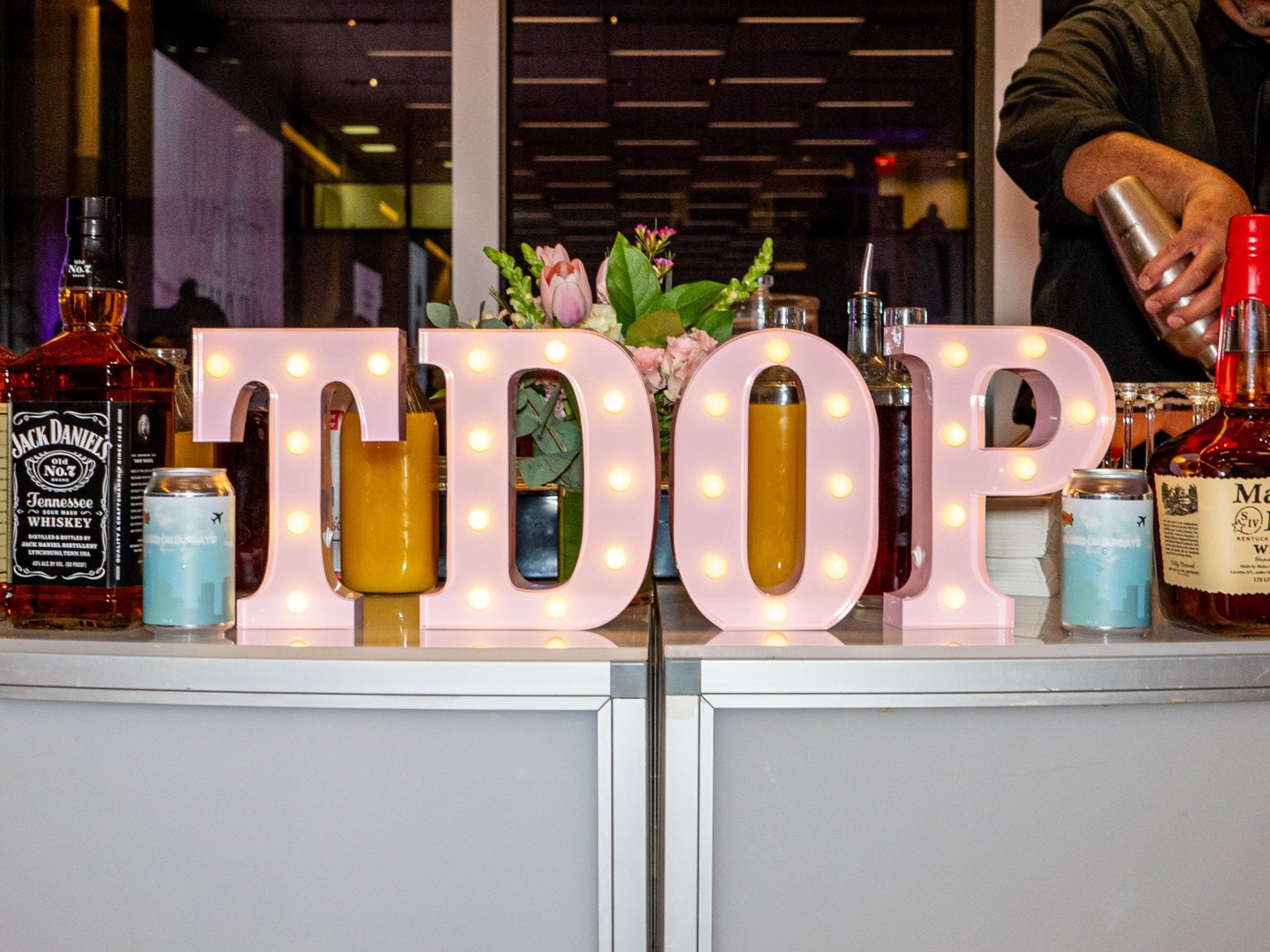 Decorative drink table with illuminated pink letters spelling 'TDOP,' bottles of liquor, a bouquet of flowers, and a bartender activating a shaker in the background.