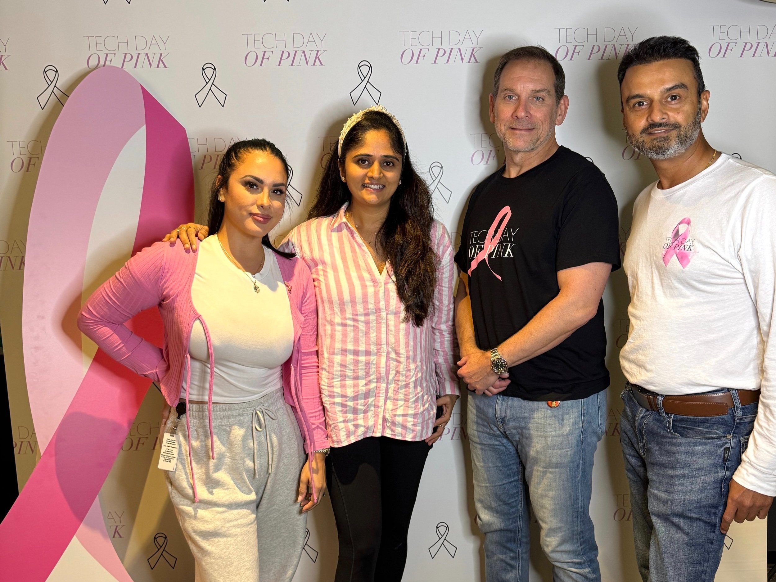 Four people standing together at a breast cancer awareness event, with a pink ribbon and a backdrop reading 'Tech Day of Pink' and pink ribbons.