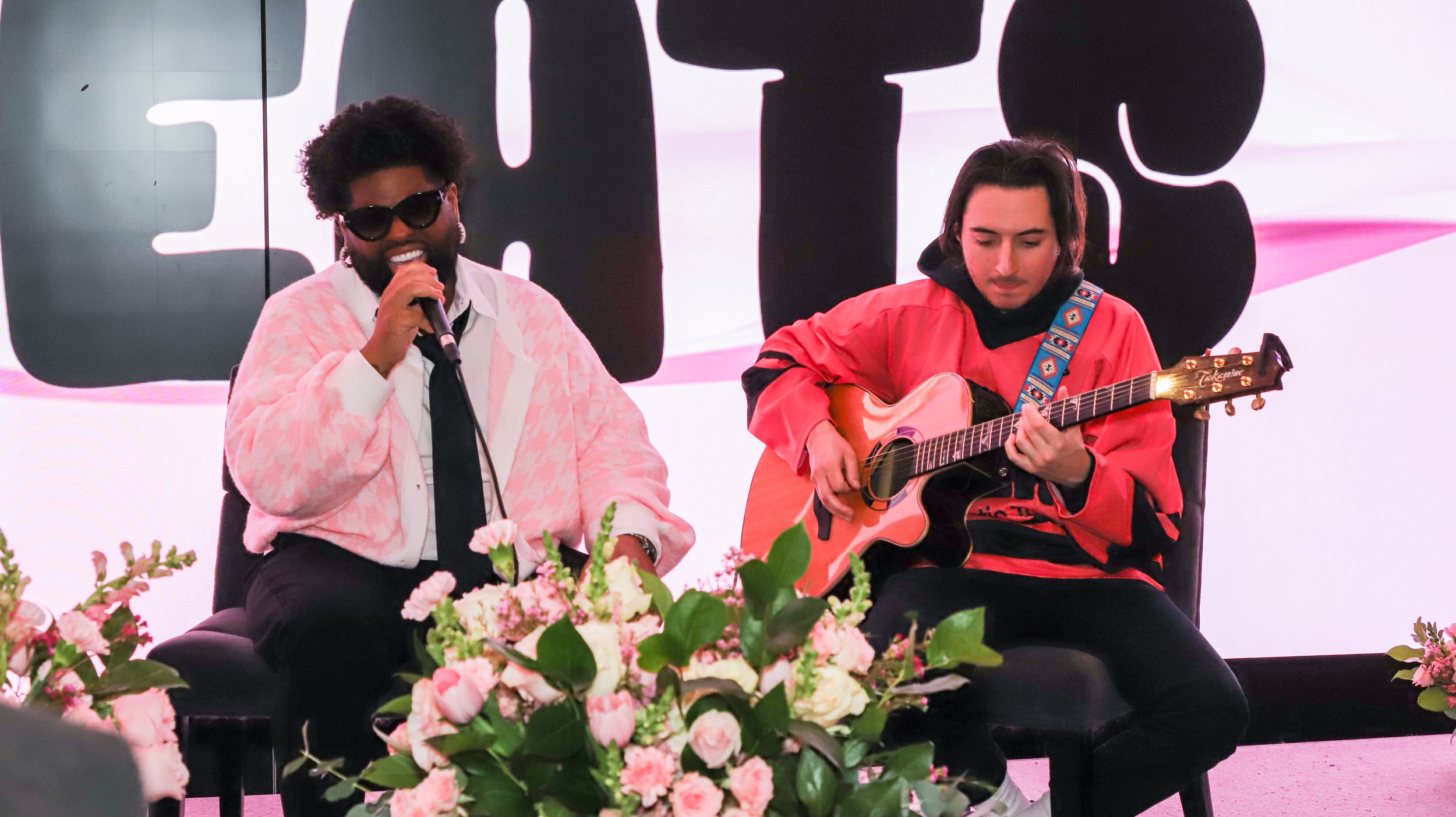 Pink Sweat$ performing on stage with a pink and black backdrop. One man is singing into a microphone, wearing sunglasses, a pink and white patterned jacket, and a white shirt with a black tie. The other man is playing an acoustic guitar, wearing a re