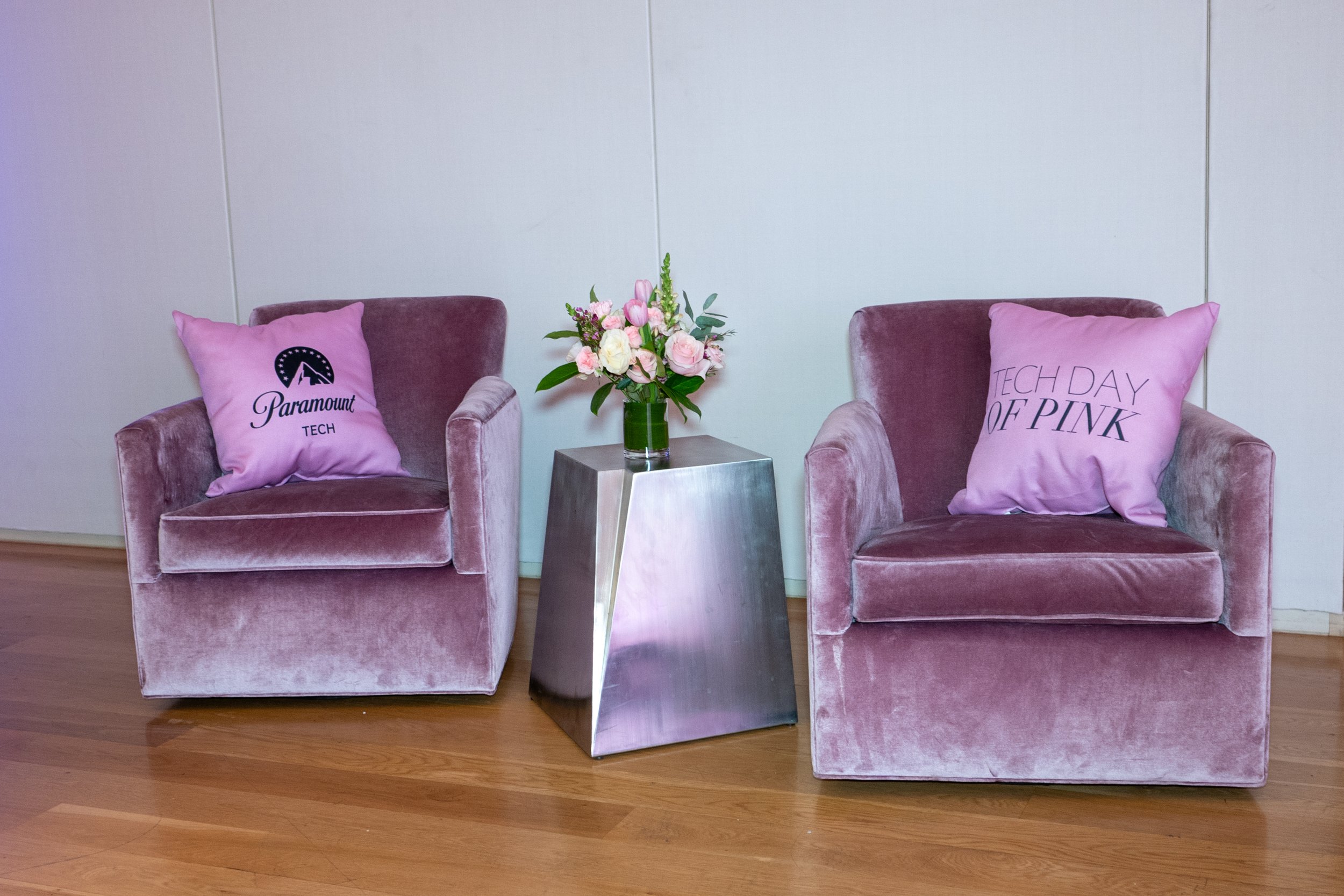 Two pink velvet armchairs with pink decorative pillows, a small metallic side table with a flower arrangement between the chairs, and a plain white wall background.