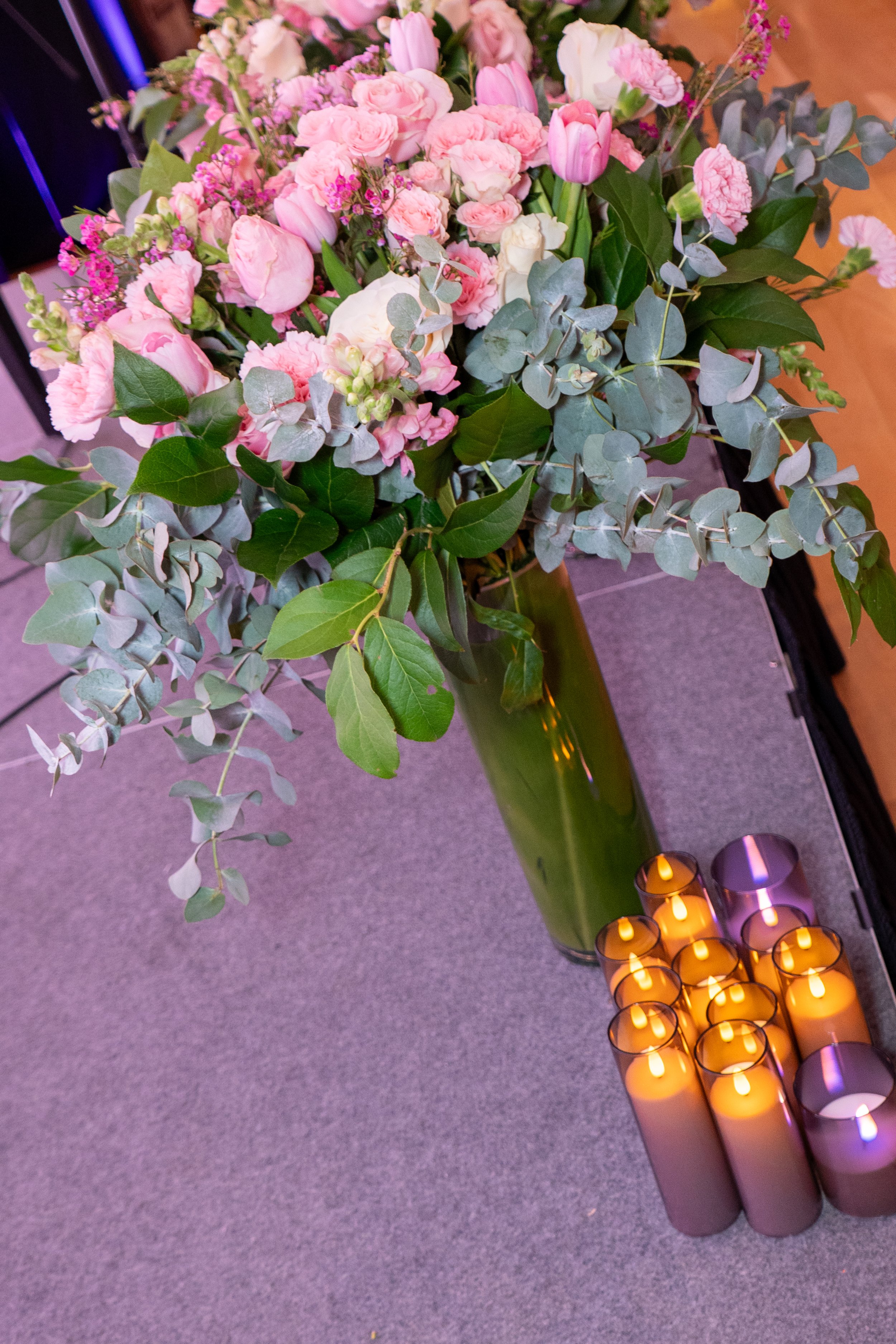 A large bouquet of pink tulips, roses, and other pink flowers with green leaves and eucalyptus in a tall green glass vase, alongside several white and purple candles on a gray carpeted surface.
