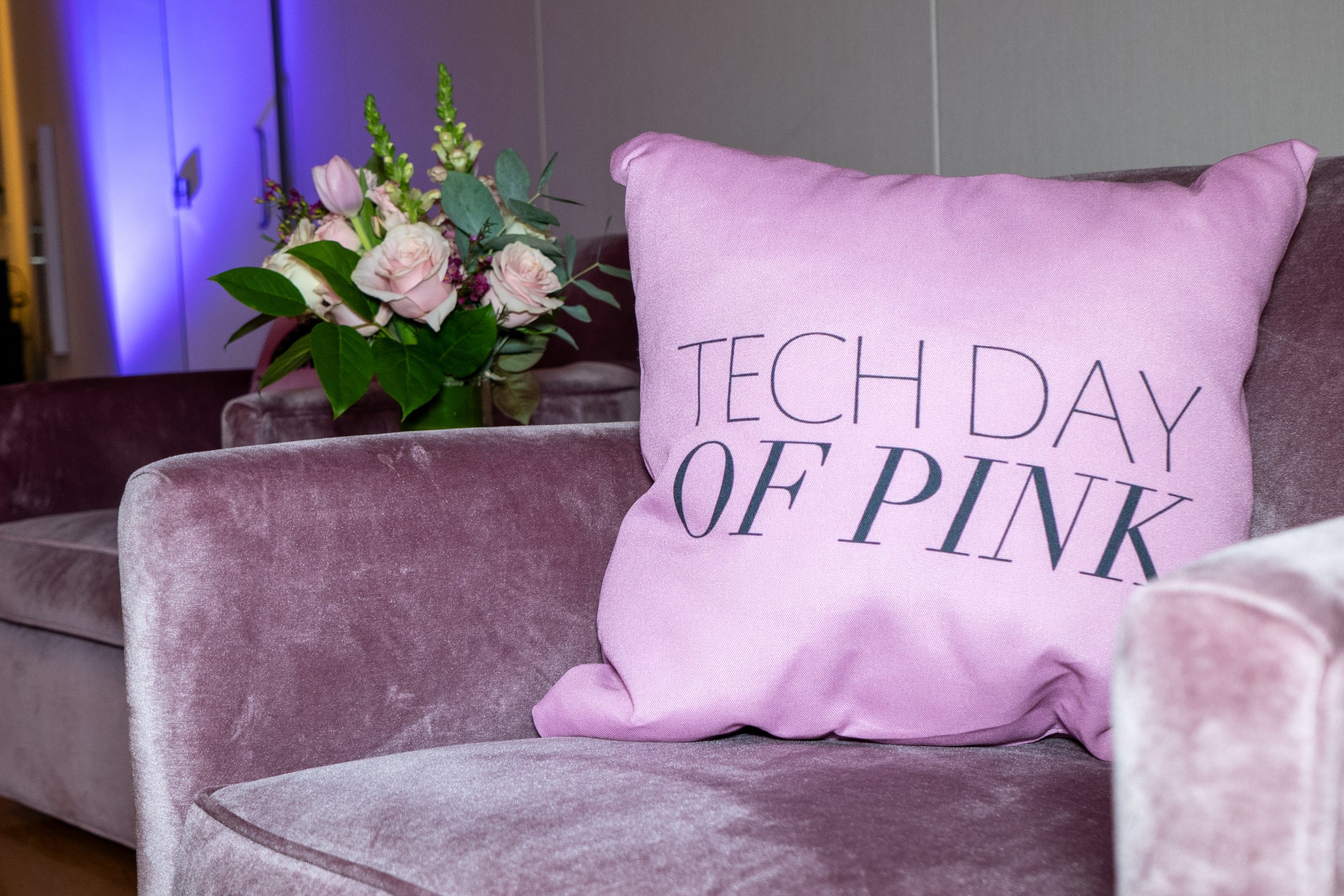 Pink velvet couch with a pink cushion that says "Tech Day of Pink" and a vase of pink flowers in the background.