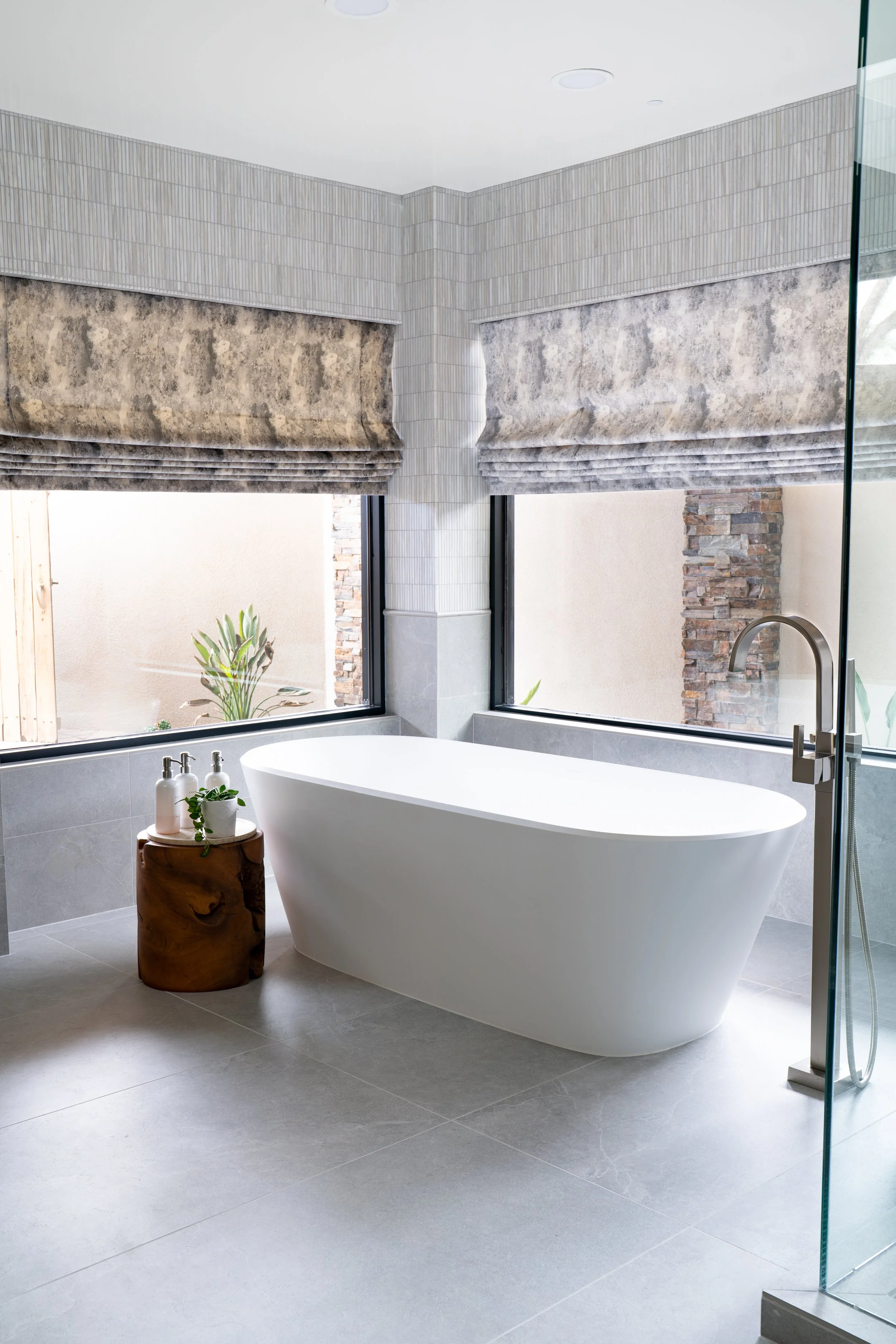 Luxury Scottsdale Paradise Valley bathroom designed by Red Egg Design Group, featuring a sculptural freestanding soaking tub, floor-mounted faucet, textured tile, custom Roman shade, natural wood accent table, and serene indoor-outdoor views.