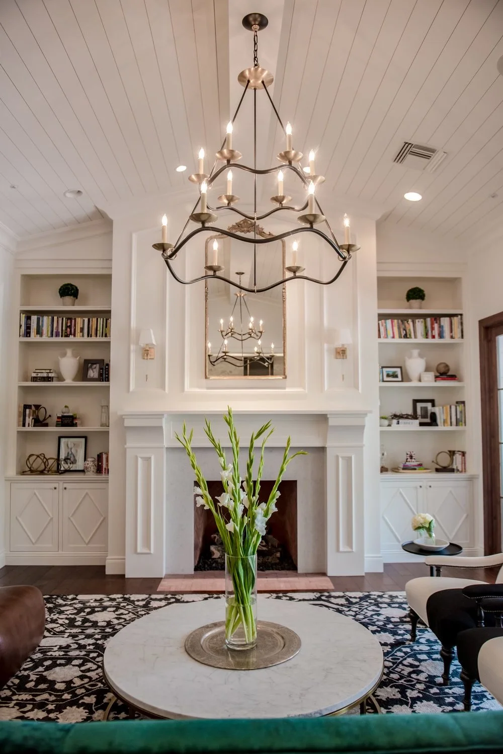 Elegant living room with white built-in bookshelves, a white marble fireplace, a large round white coffee table with a gold tray and glass vase of white flowers, and a black and white patterned rug. A chandelier with multiple candle-style bulbs hangs