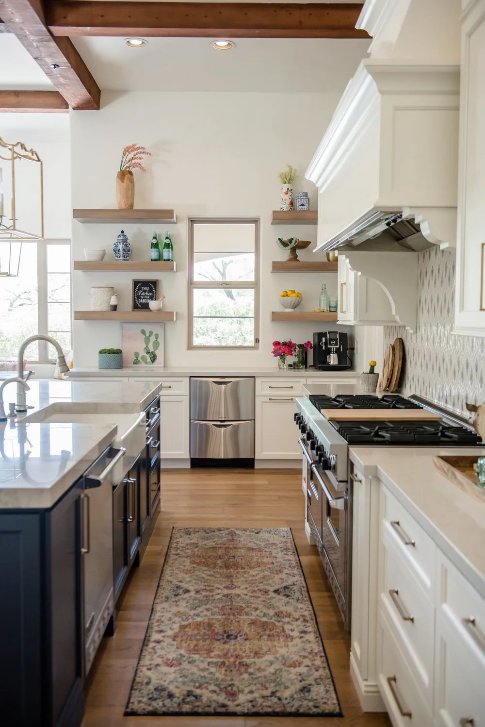 A bright, modern kitchen with white cabinets, open shelves, a stainless steel dishwasher, gas stove, and wooden flooring. Decor includes plants, bowls, and decorative bottles, with a small window and a decorative rug on the floor.
