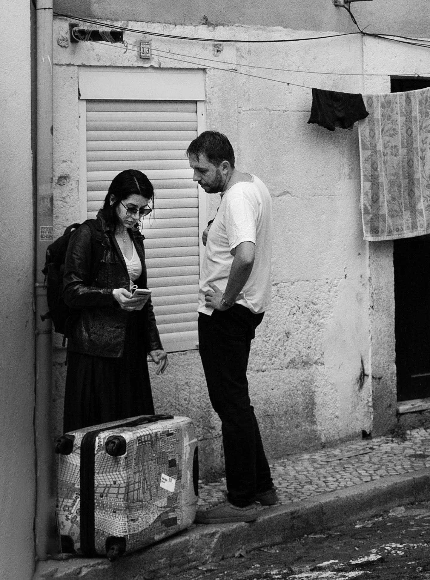 To personer står på en fortovskant foran en gammel væg, med et stykke bagage på jorden foran dem, de ser på en mobiltelefon. 
