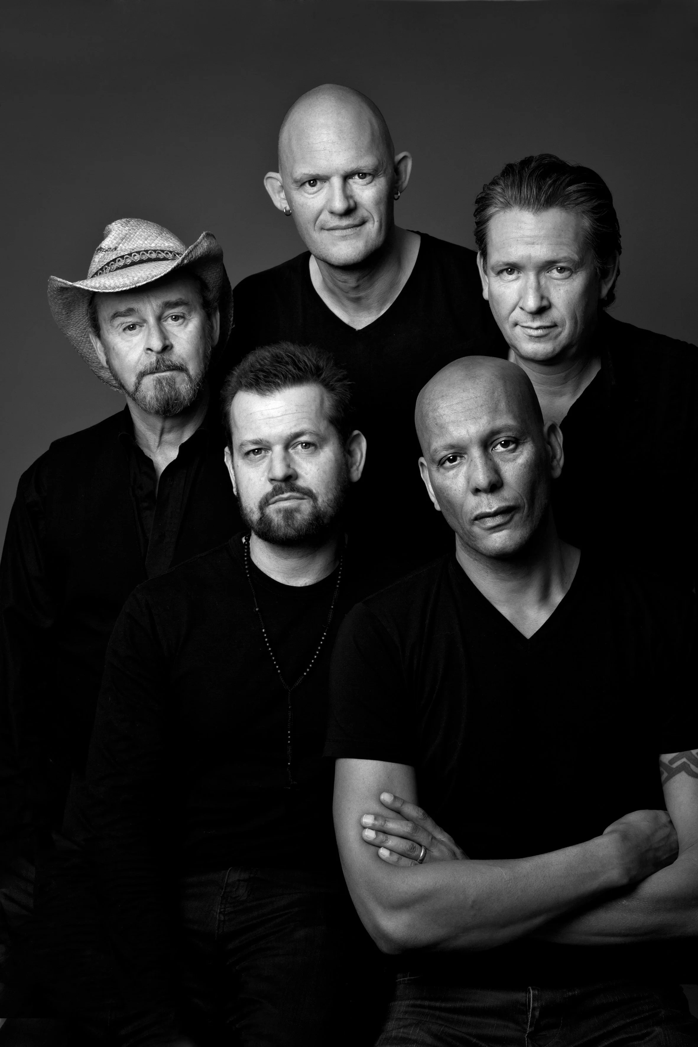 Sort-hvid gruppebillede af seks mænd, nogle med beard, en med hatte, alle i sorte t-shirts, portræt med neutral baggrund.