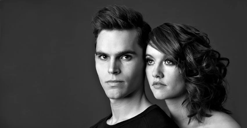 Sort-hvid portrætfotografi af en ung mand og kvinde, der står tæt sammen mod en mørk baggrund.