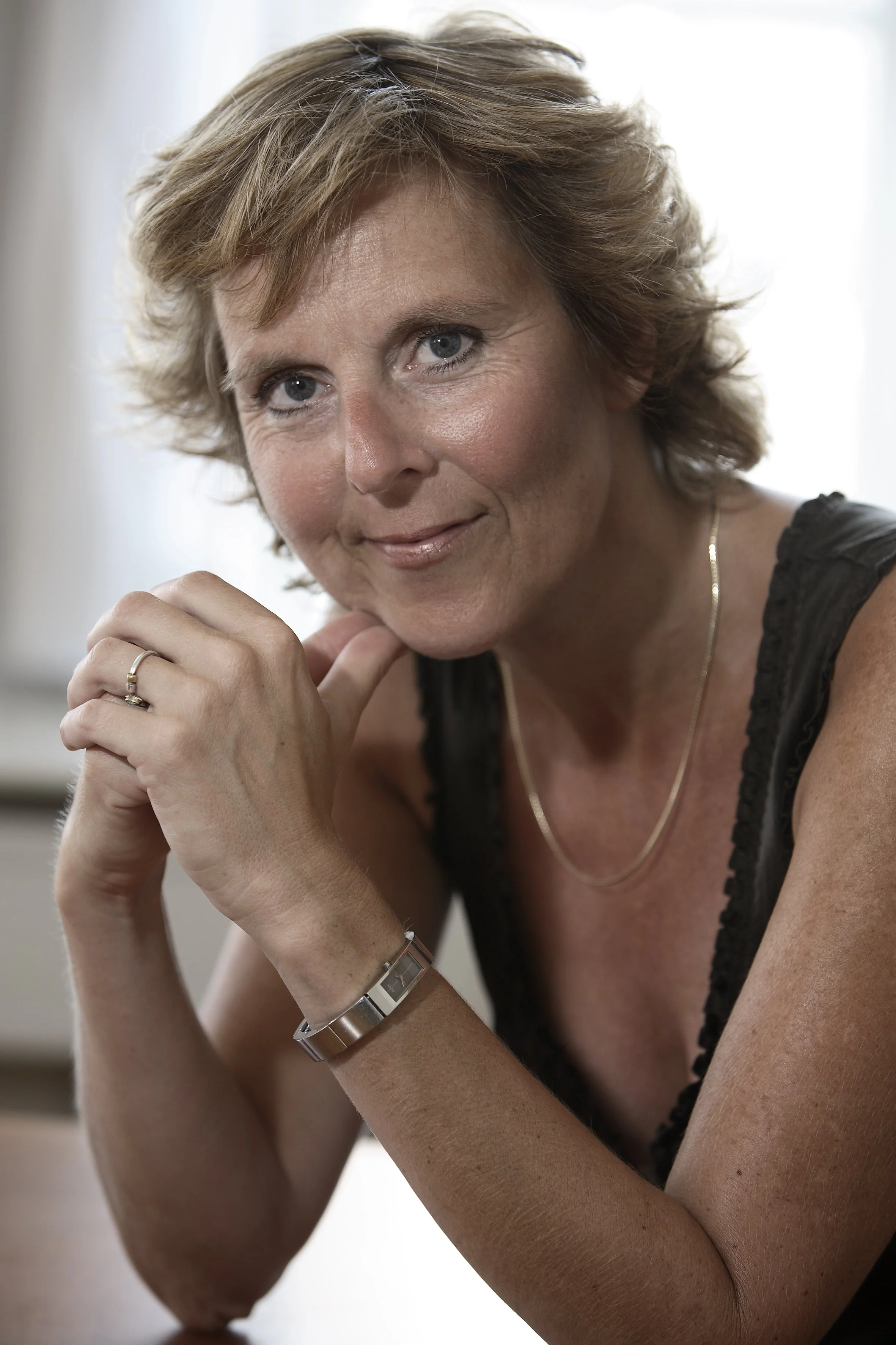 Portræt af en midaldrende kvinde med lyst, kort hår, blå øjne, iført sort ærmeløst top, guld halskæde, armbånd og ring, ser ind i kameraet med et blidt smil.