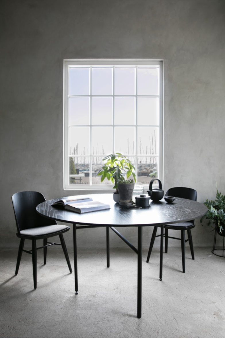 Elegant spisebord med tre sorte stole, en grøn plante i midten, en åben bog, en sort kobberkande og små skåle, i et minimalistisk rum med stor vindue, grå væg og cementgulv.