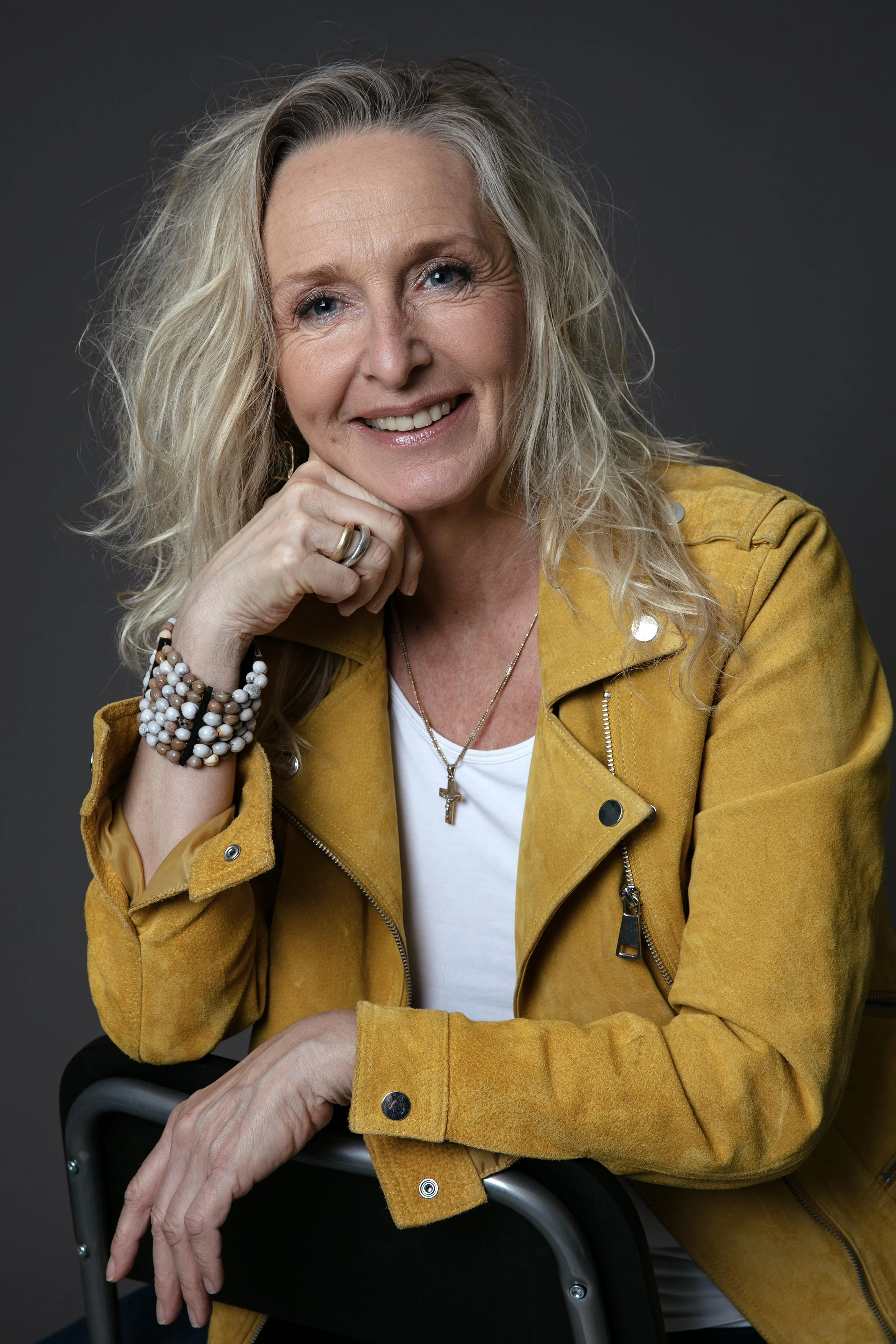 En smilende ældre kvinde med blond, wavy hår, iført en gul jakke, hvide t-shirt, smykker og et armbånd, sidder og holder i en sort stol mod en grå baggrund.