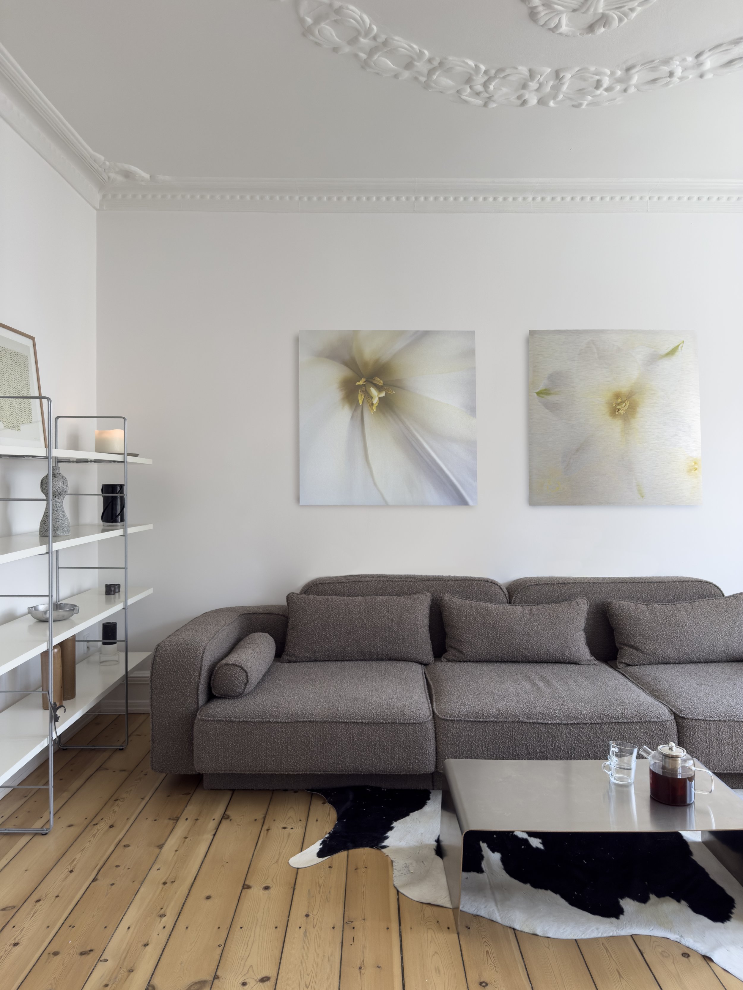 Et stueindretning med en beige hjørnesofa under to delikate blomsterbilleder på væggen, en metalhylde med dekorative genstande til venstre, og et coffee table med glasvase og the eller kaffe i midten. Gulv i træ og sort-hvid tæppe.