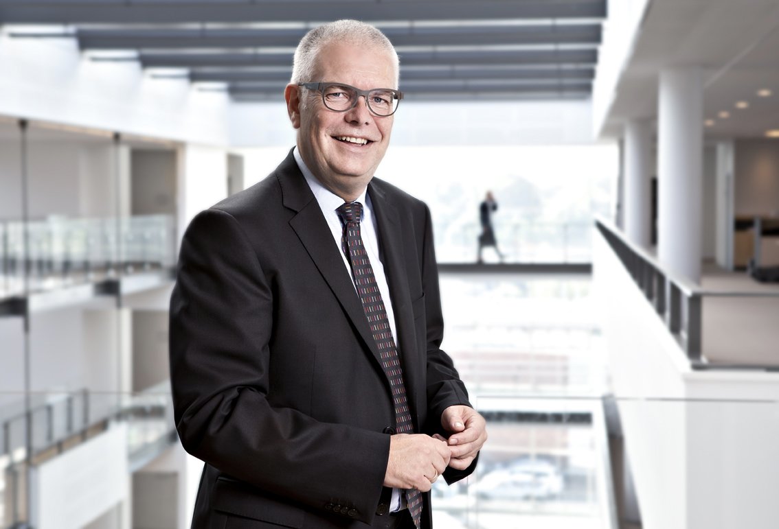 En mand med gråt hår og briller iført sort jakkesæt, hvid skjorte og slips står i en moderne kontorbygning.