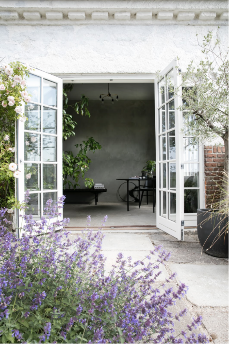 Åbent dørparti til en privat have eller terrasse med blomster og planter udenfor, indendørs et minimalistisk rum med et bord og stole.