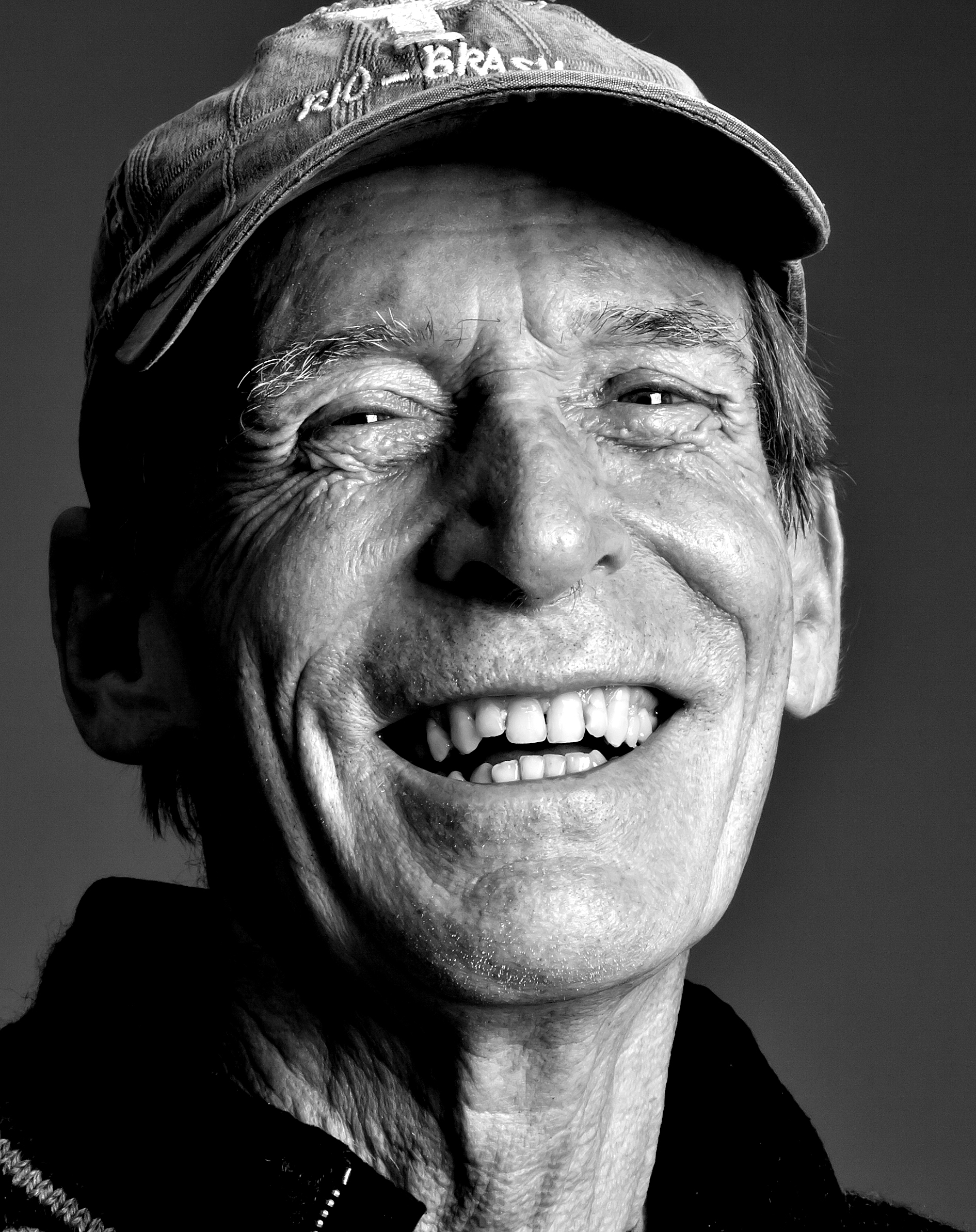 Sort-hvid portræt af en ældre mand iført kasket, smilende med synlige tænder, under et klart lys.