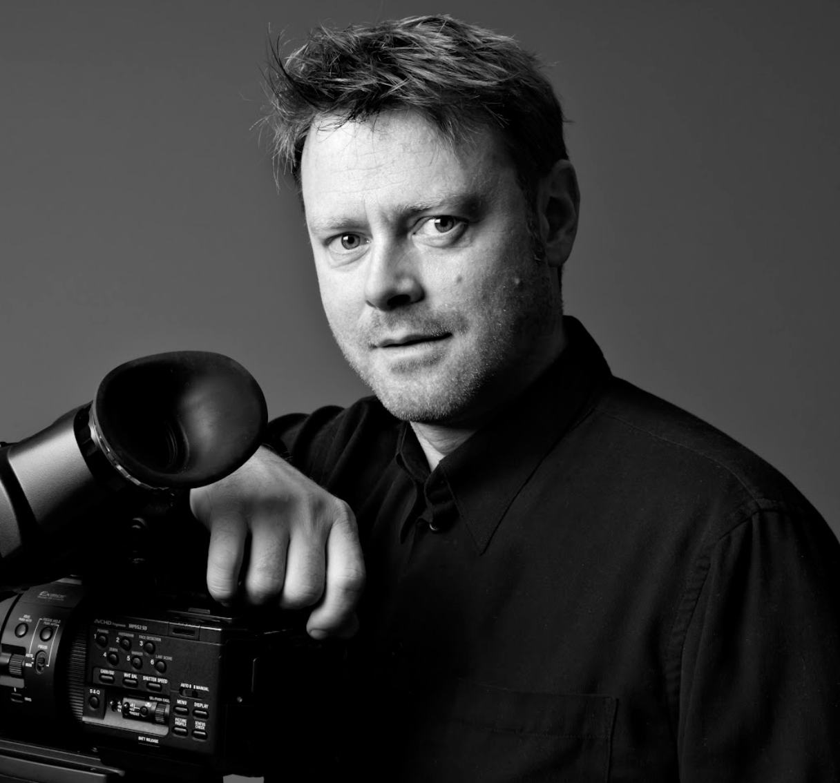 Sort-hvid portræt af fotograf Rasmus Aude, der holder et professionelt videokamera.