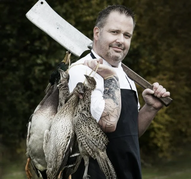 Man med tatoveringer og forklæde, der bærer en kødøkse, med flere fugle hængende på hans skulder.
