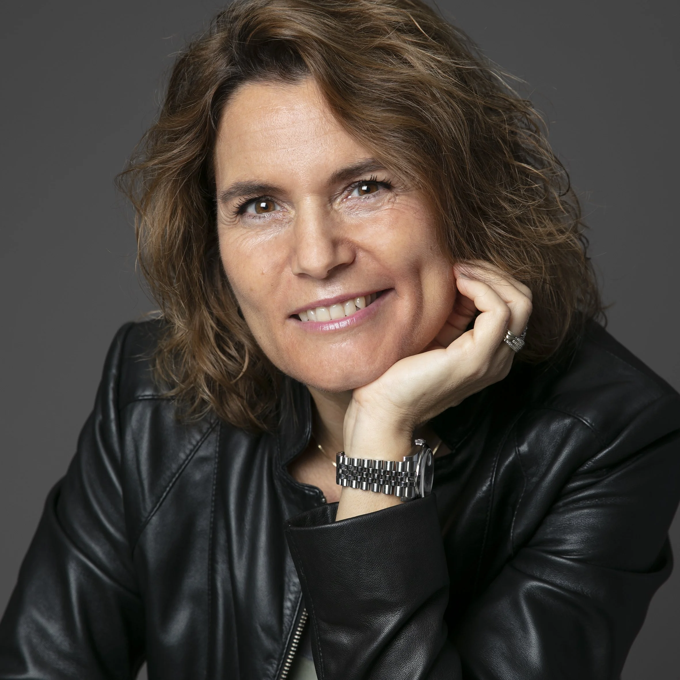 Portræt af en smilende kvinde med brunt, gråt og blond hår, iført sort læderjakke og sølv armbånd, mod en grå baggrund.