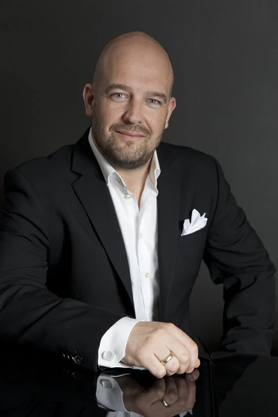 Portræt af en mand i sort jakkesæt med hvid skjorte, sidder ved et bord mod en mørk baggrund.