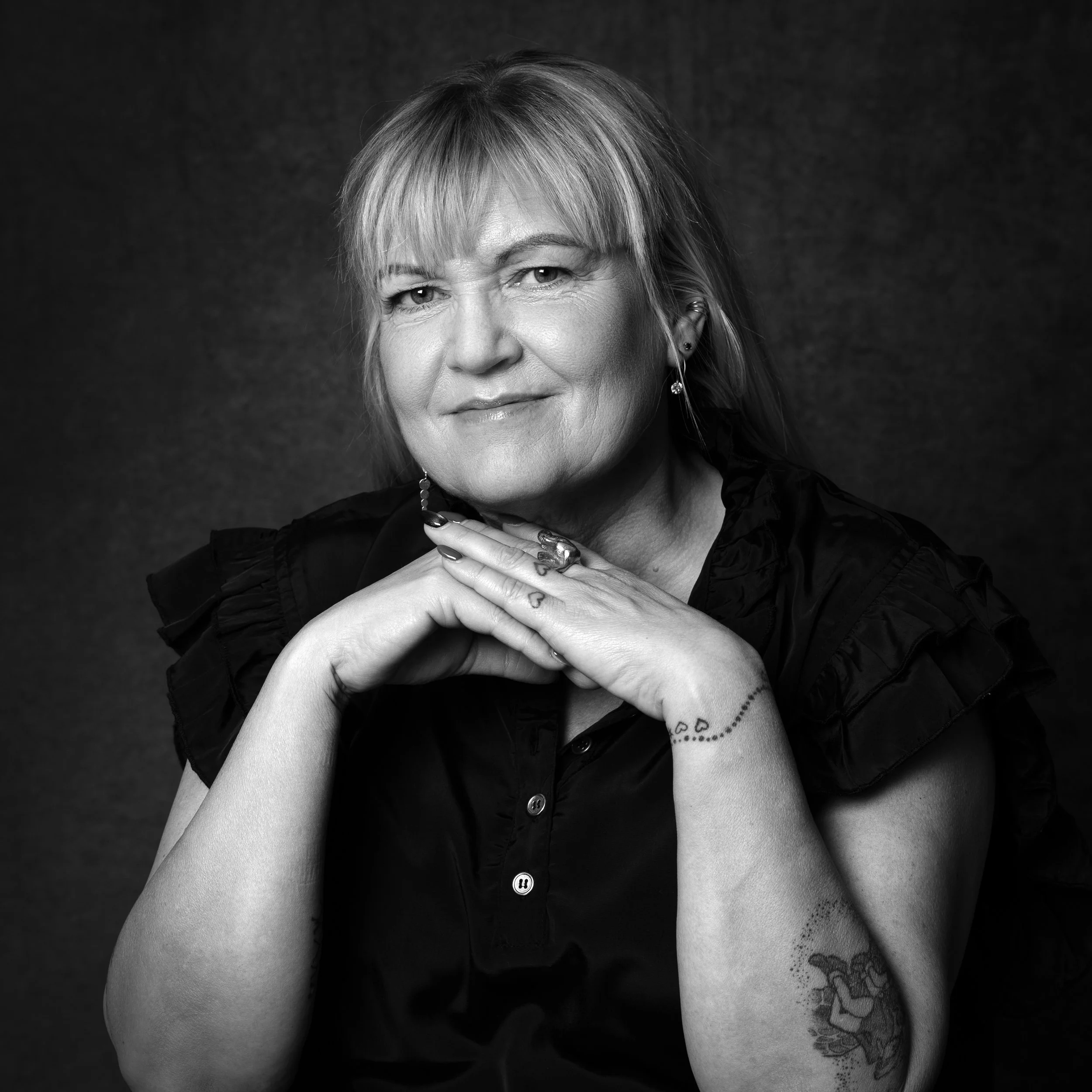 Sort-hvid portræt af en ældre kvinde med blondt hår, smilende, iført sort tøj med rynkefri ansigt, tatoveringer på hænder og arme, armene krydset foran kinnen.
