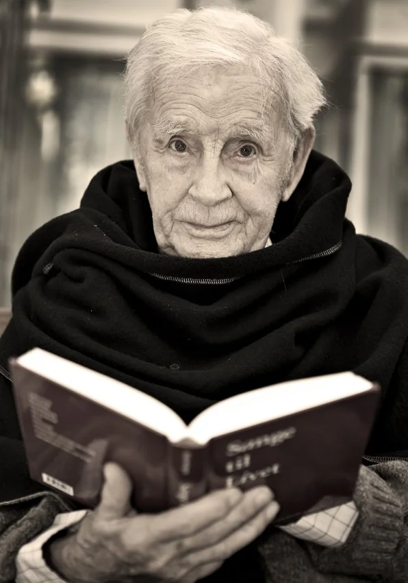 Ældre person med hvidt hår, iført sort tøj, læsende en bog.