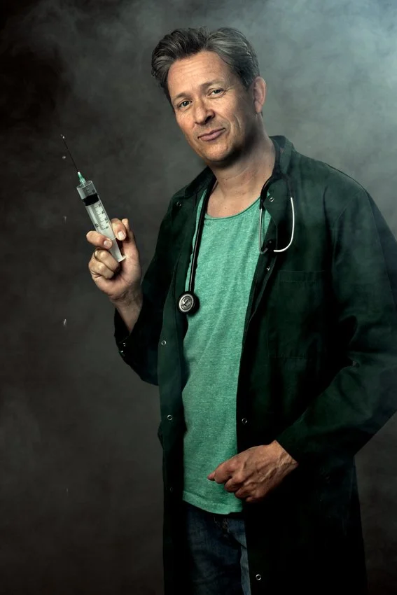 En mand iført en grøn T-shirt og en mørk grøn jakke, holder en sprøjte i hånden, med en baggrund i grå nuancer.