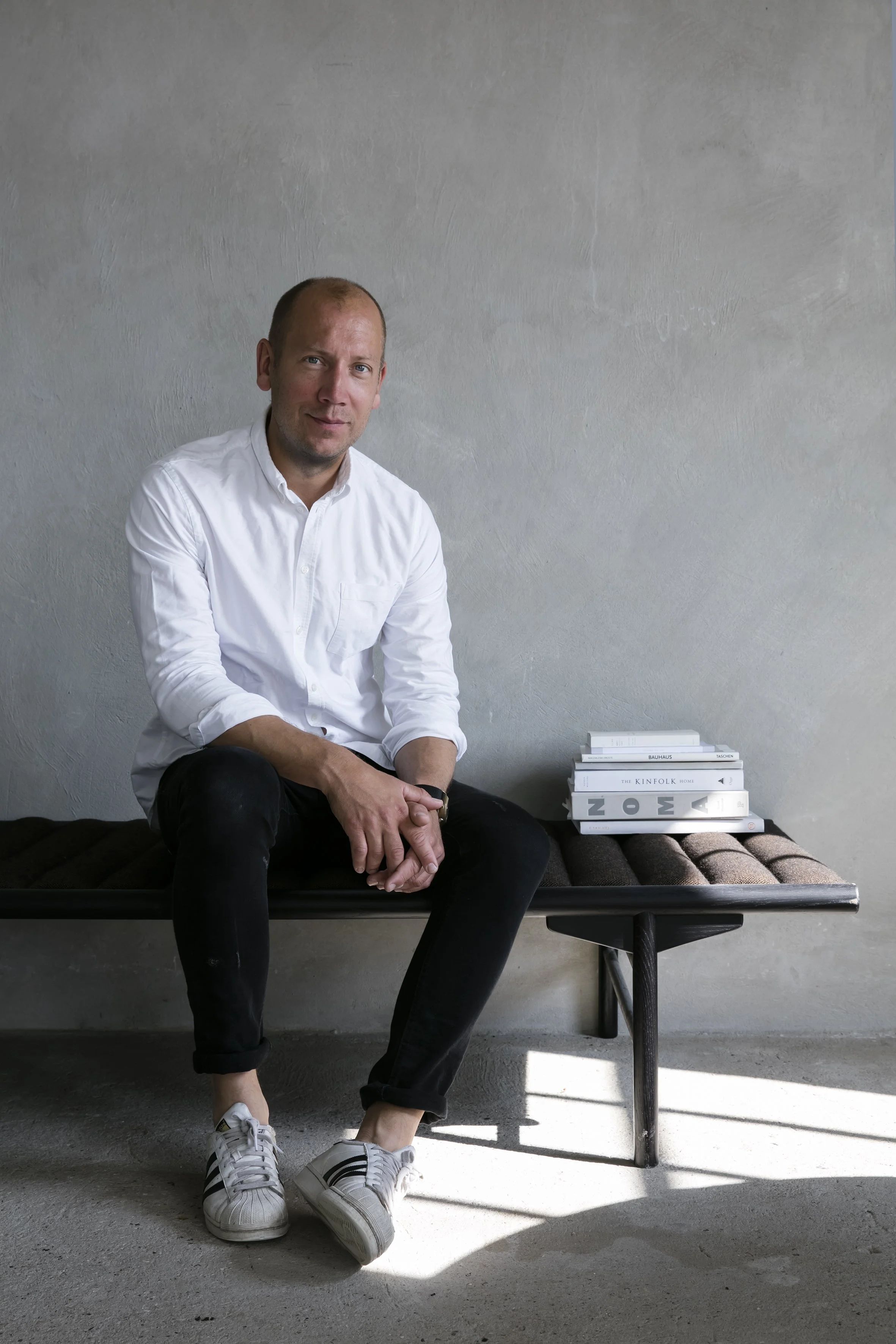 En mand i hvid skjorte og sorte bukser sidder på en sort træbænk med en bunke bøger ved siden af.