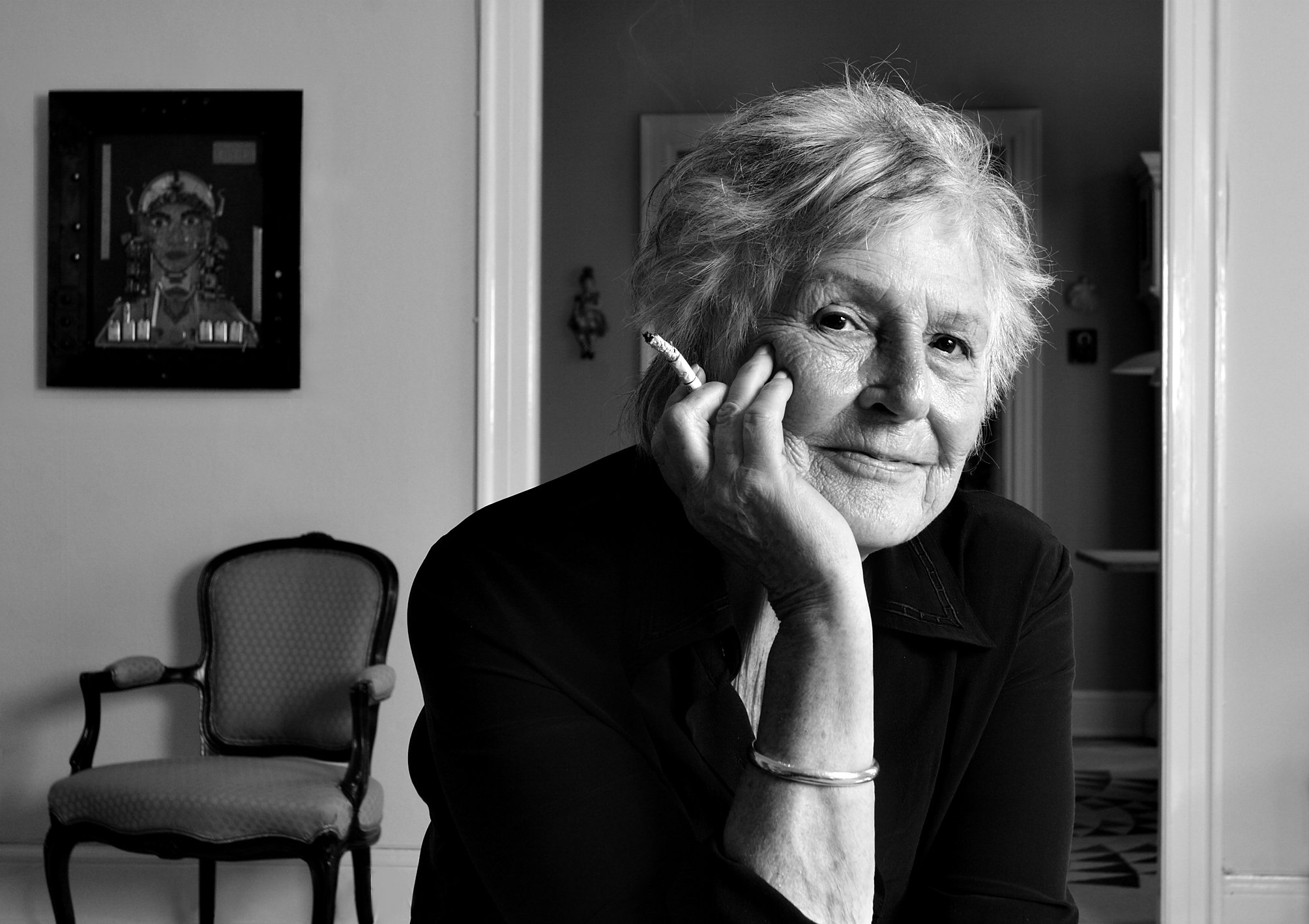 Sort-hvid foto af en ældre kvinde, der sidder med en cigar i hånden, med en hyggelig indretning i baggrunden.