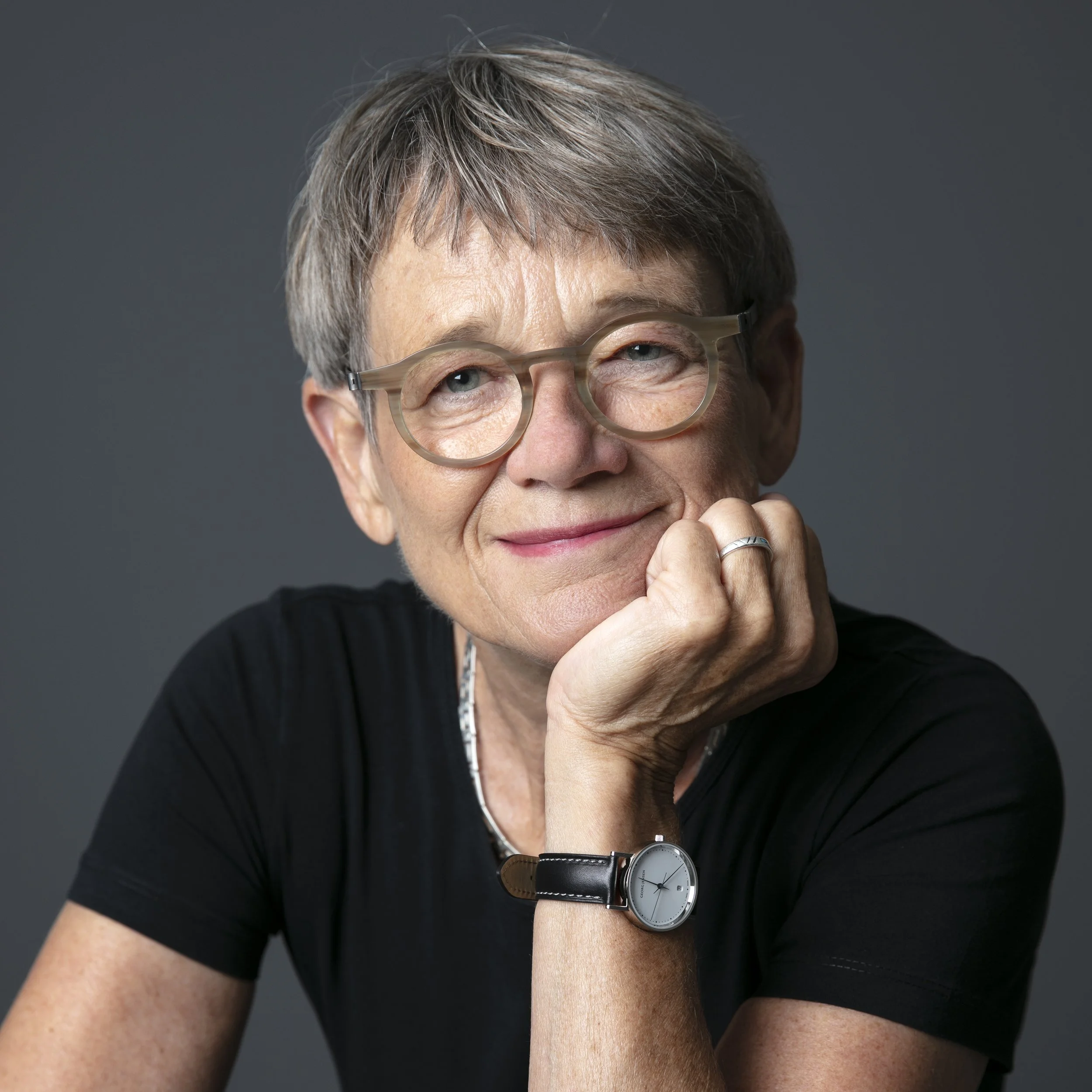 Et portræt af en glad ældre kvinde med kort gråt hår, briller, og et sort t-shirt, der hviler sit hoved på sin hånd med et smil.