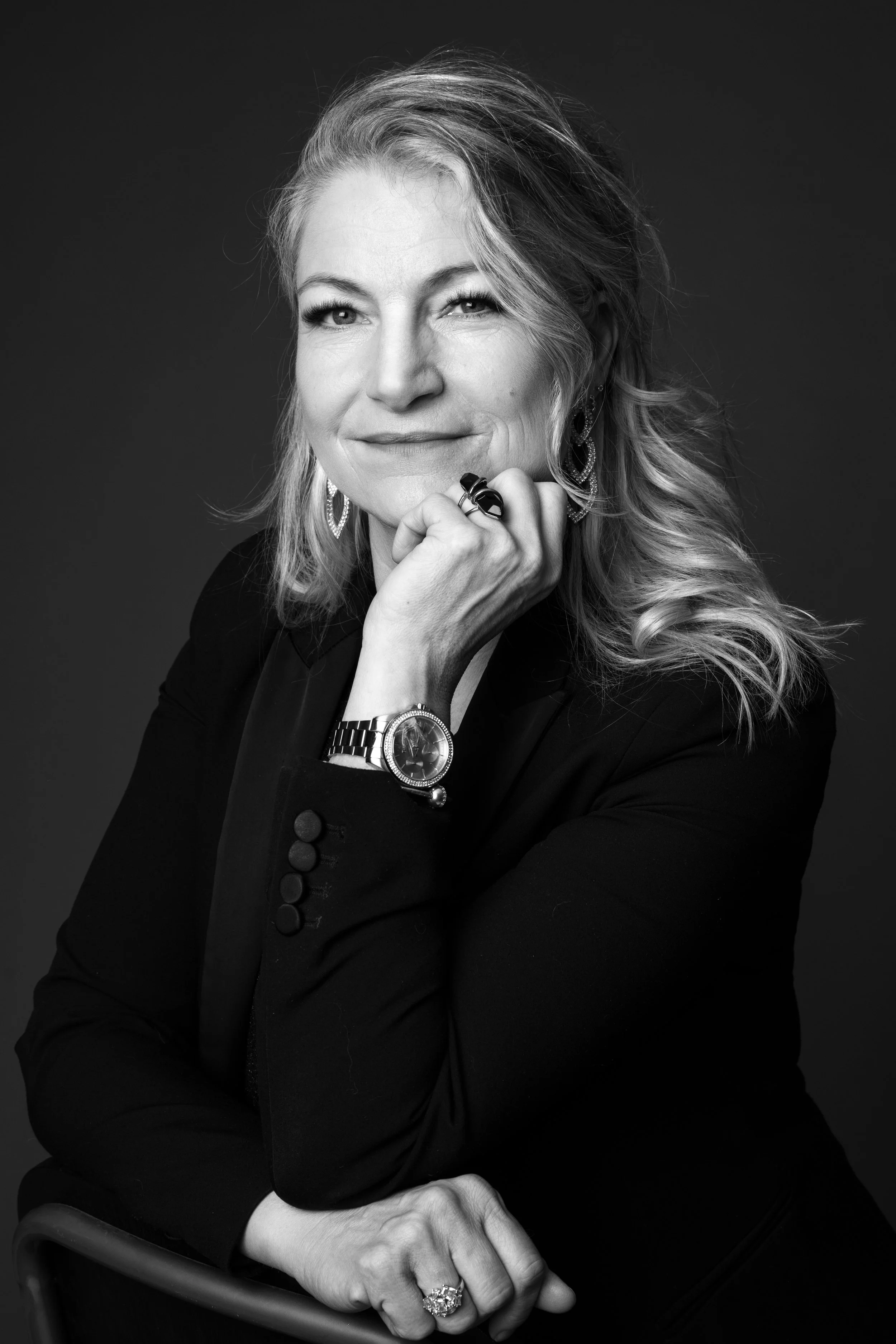Sort-hvid portrætfotografi af en kvinde med blondt, bølget hår, iført en sort blazer og smykker, smiling let, med hånden under hagen, mod en uklar sort baggrund.