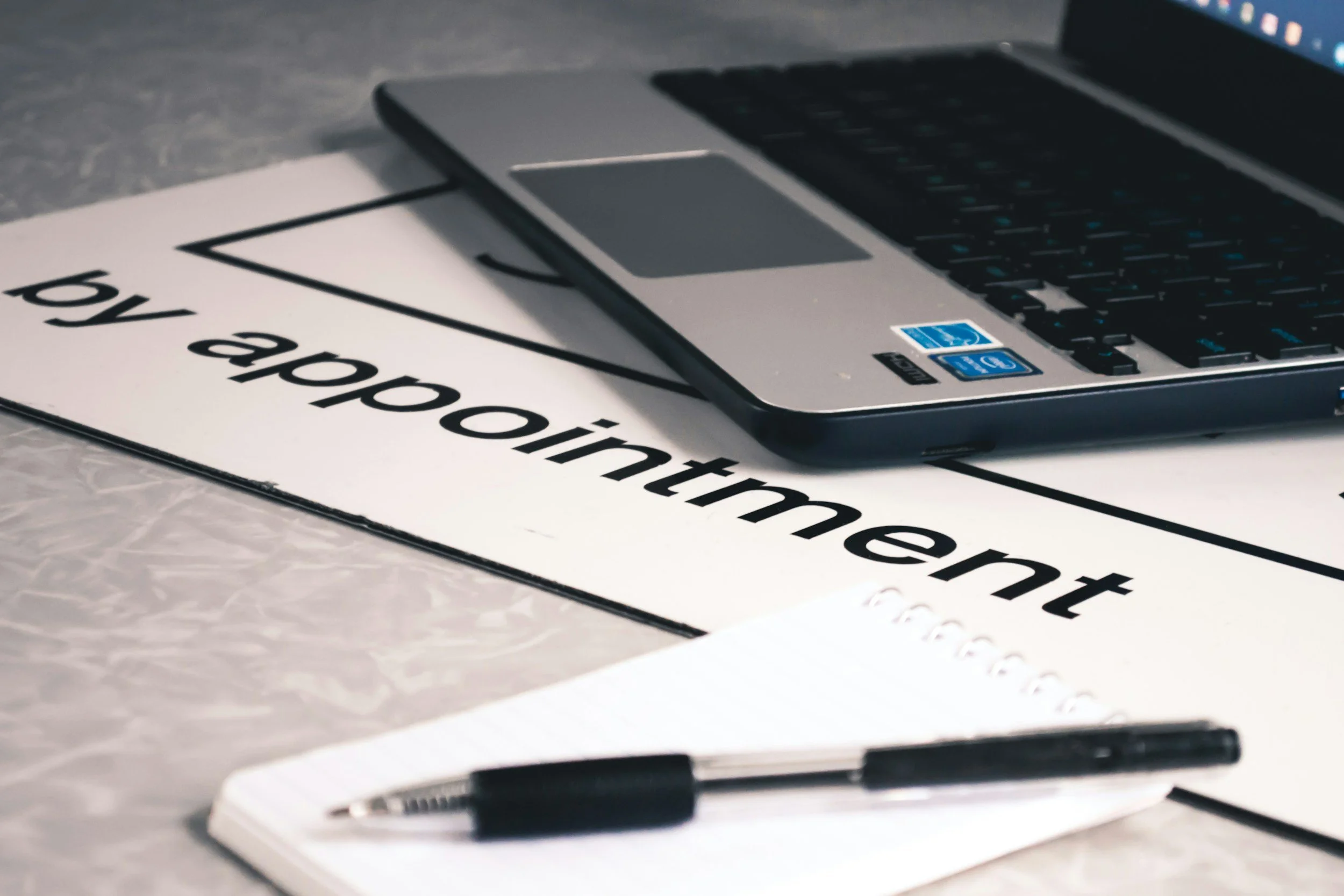 A laptop, a notebook with a pen, and a sign that says 'by appointment' on a marble surface.