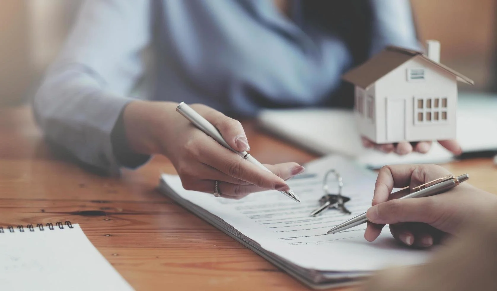 Two people exchanging documents, one holding a small model house, on a wooden desk.