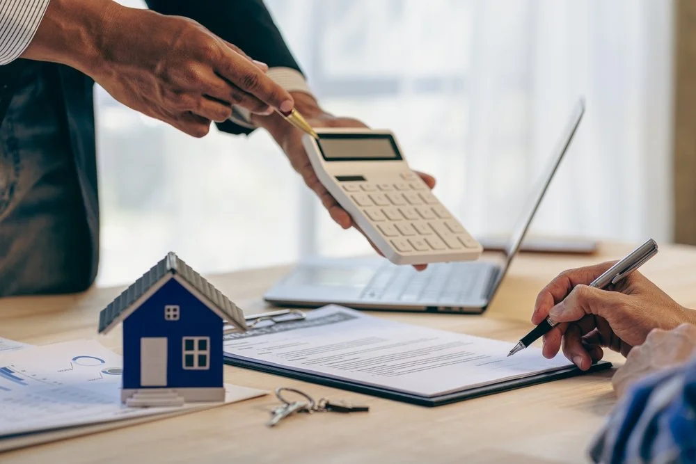 Two people discussing a real estate deal, one holding a calculator and pointing at it with a pen, the other taking notes with a pen; a small model house, keys, and documents are on the table.