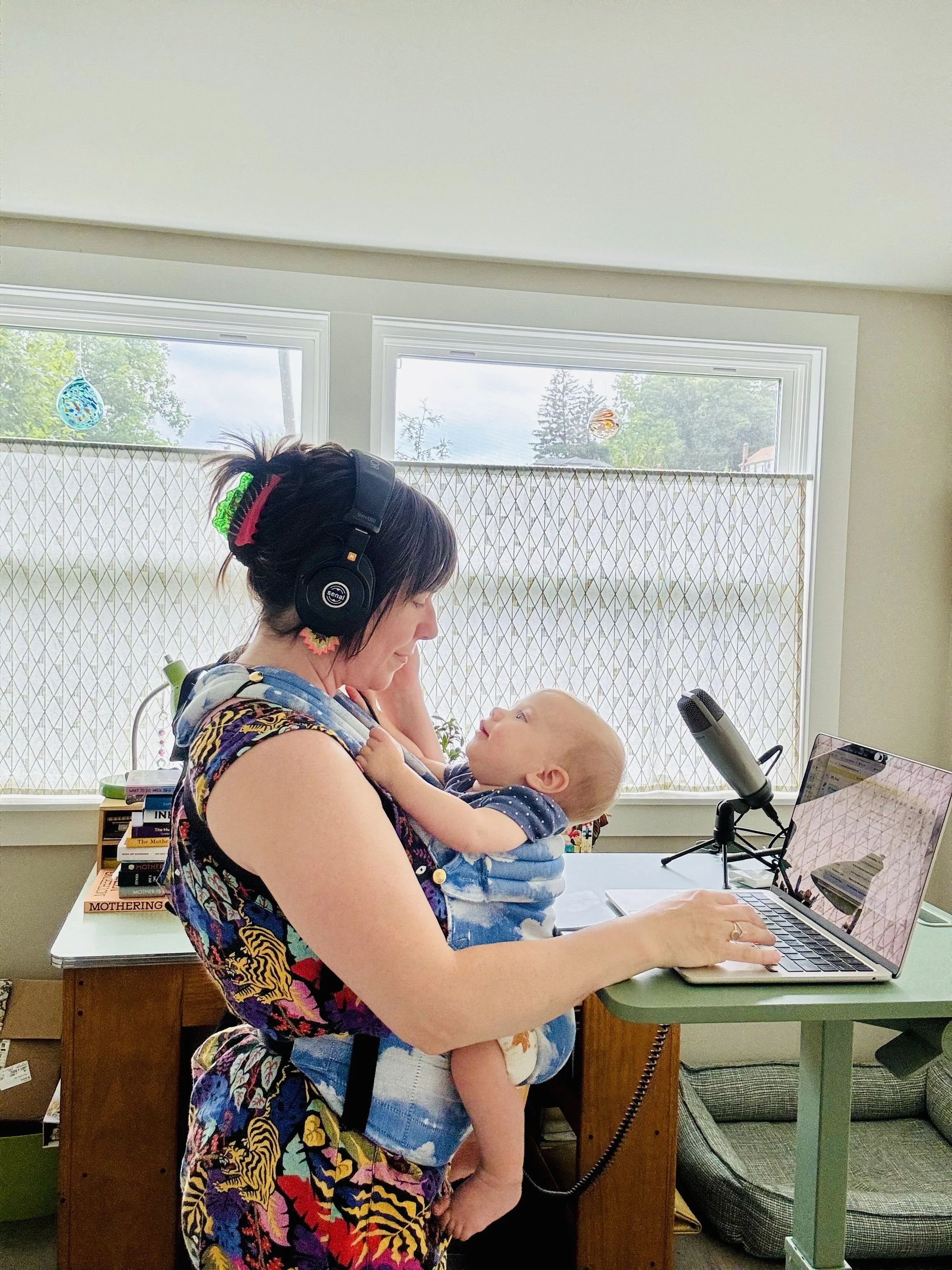 A woman with dark hair, wearing large black headphones and a colorful, patterned top, holding a young child in her arms. The woman is seated at a desk with a laptop, in a room with large windows and a bookshelf in the background.