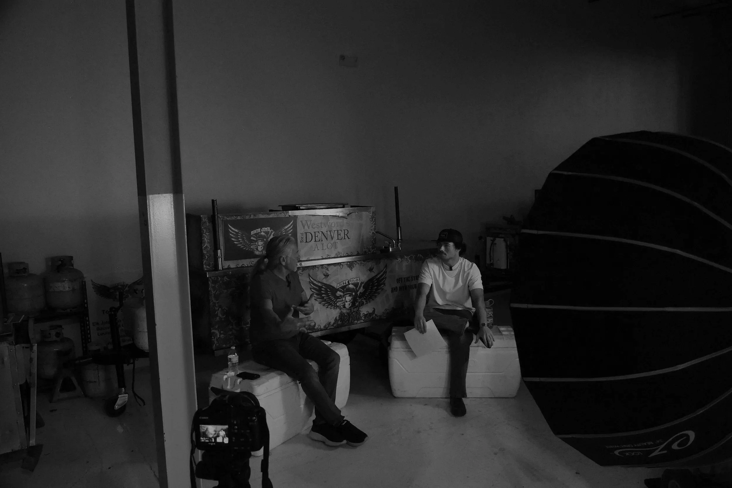 Two people sitting and talking in a dimly lit indoor setting, with a large decorative hot air balloon sculpture on the right and a camera on a tripod in the foreground.