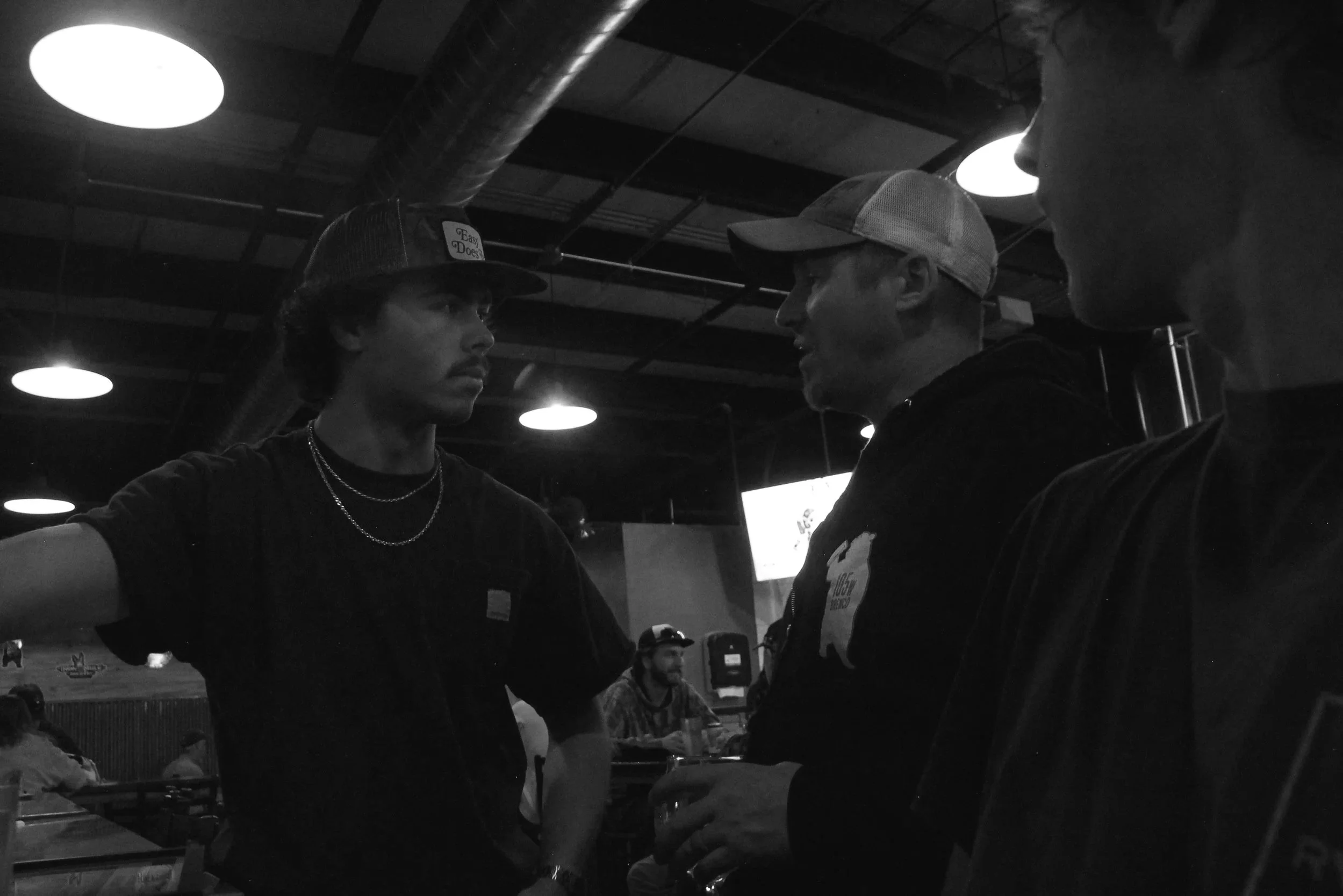 Three men are engaged in a conversation in a dimly lit indoor setting, likely a restaurant or bar, with more people and tables visible in the background.