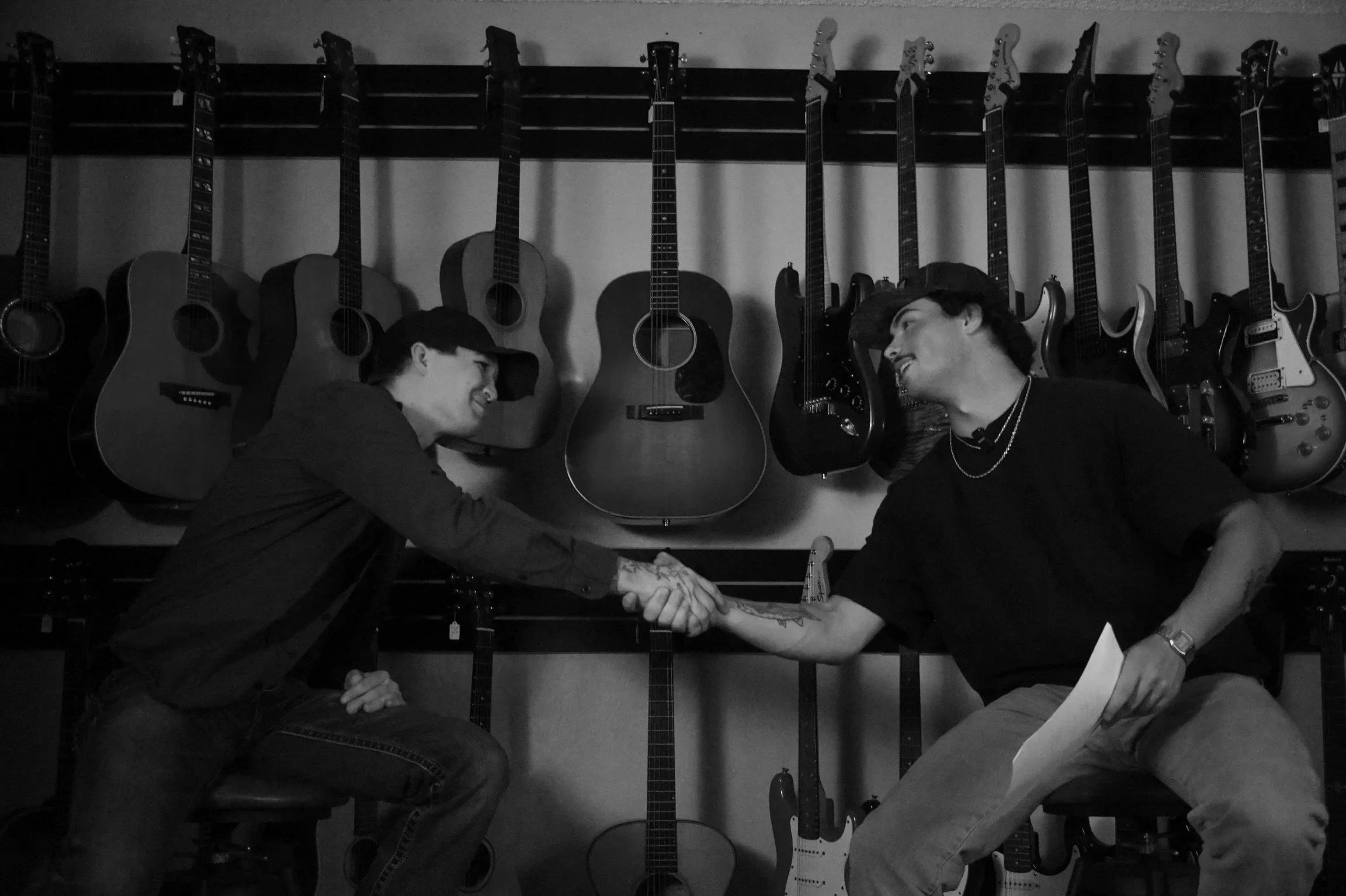Two people shaking hands in a guitar shop, with guitars hanging on the wall behind them.