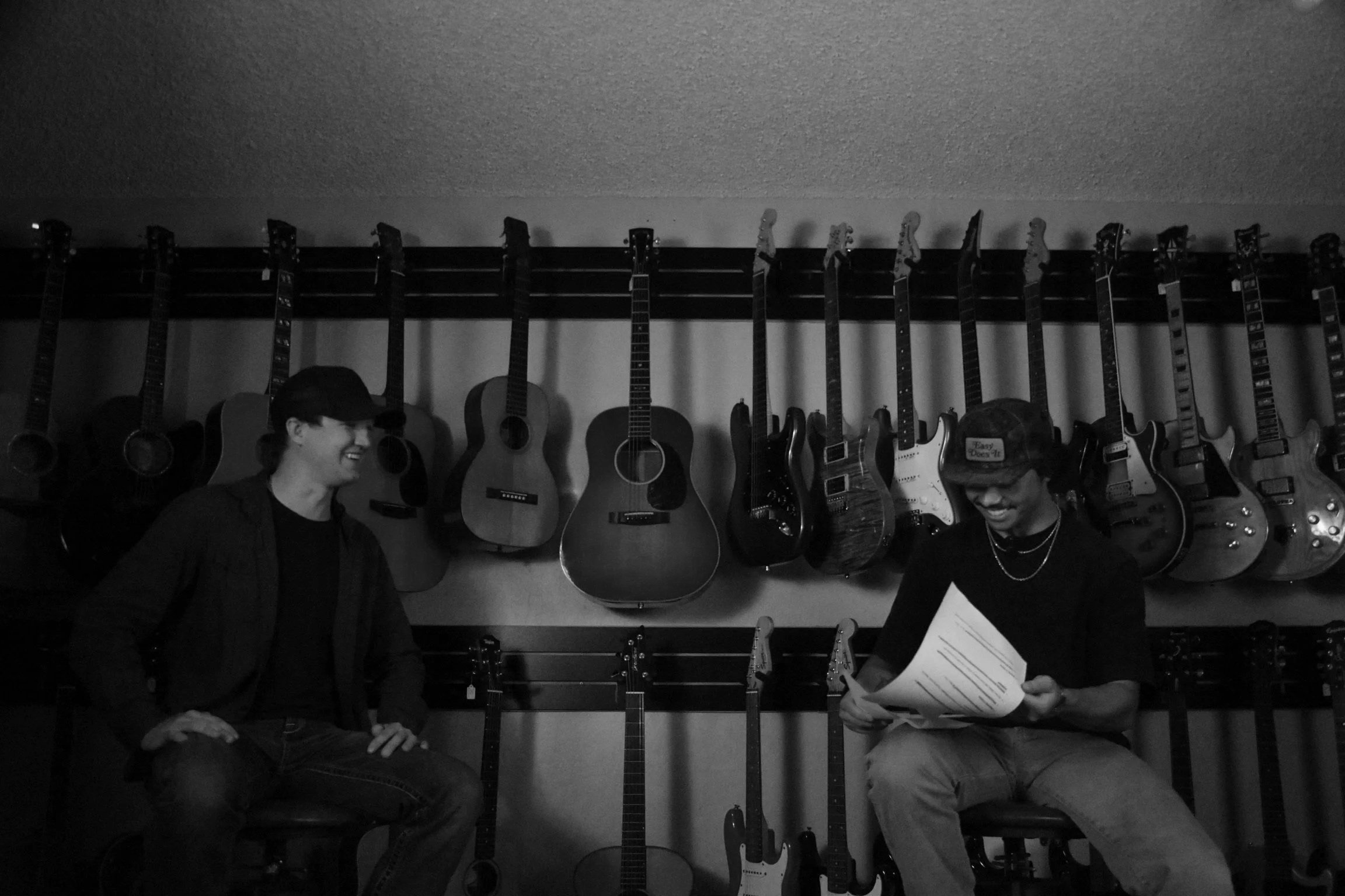 Two young men sitting in a music store with guitars hanging on the wall behind them, one smiling and the other reading a paper.