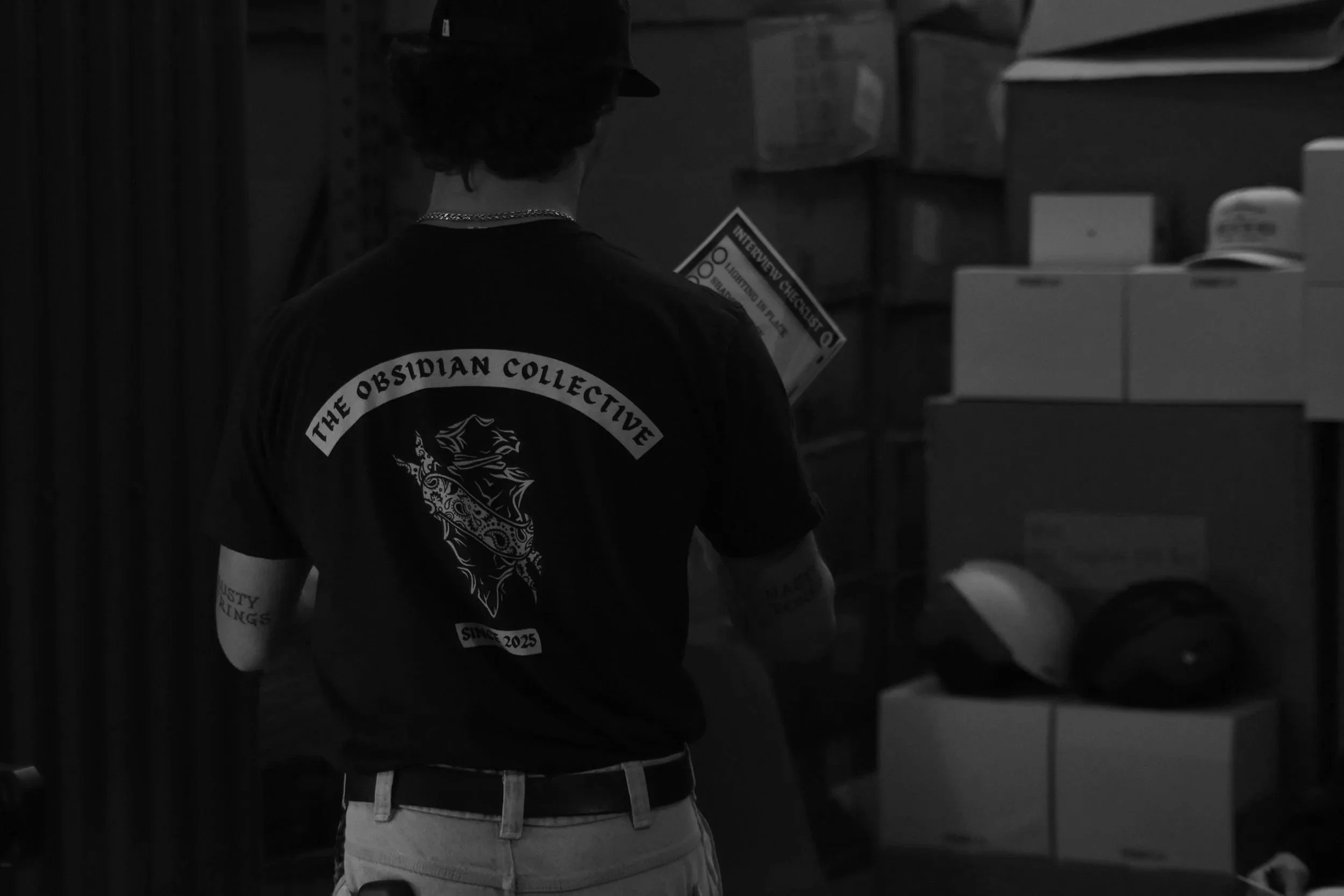 Person wearing a T-shirt with the text 'The Obsidian Collective' printed on the back, standing in a storage room with boxes and helmets.