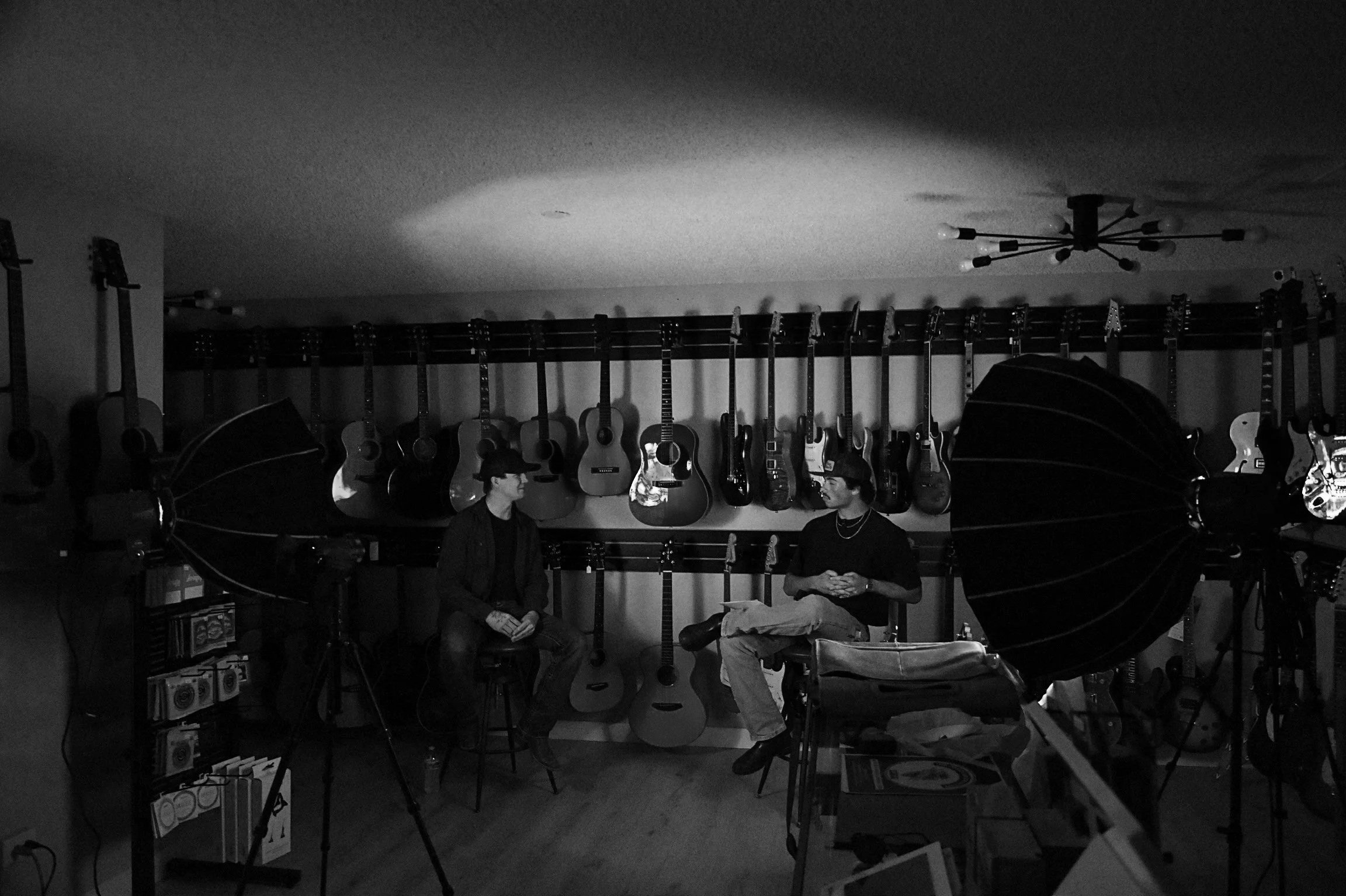 Two people sitting in a music store surrounded by guitars hanging on the wall and studio equipment.