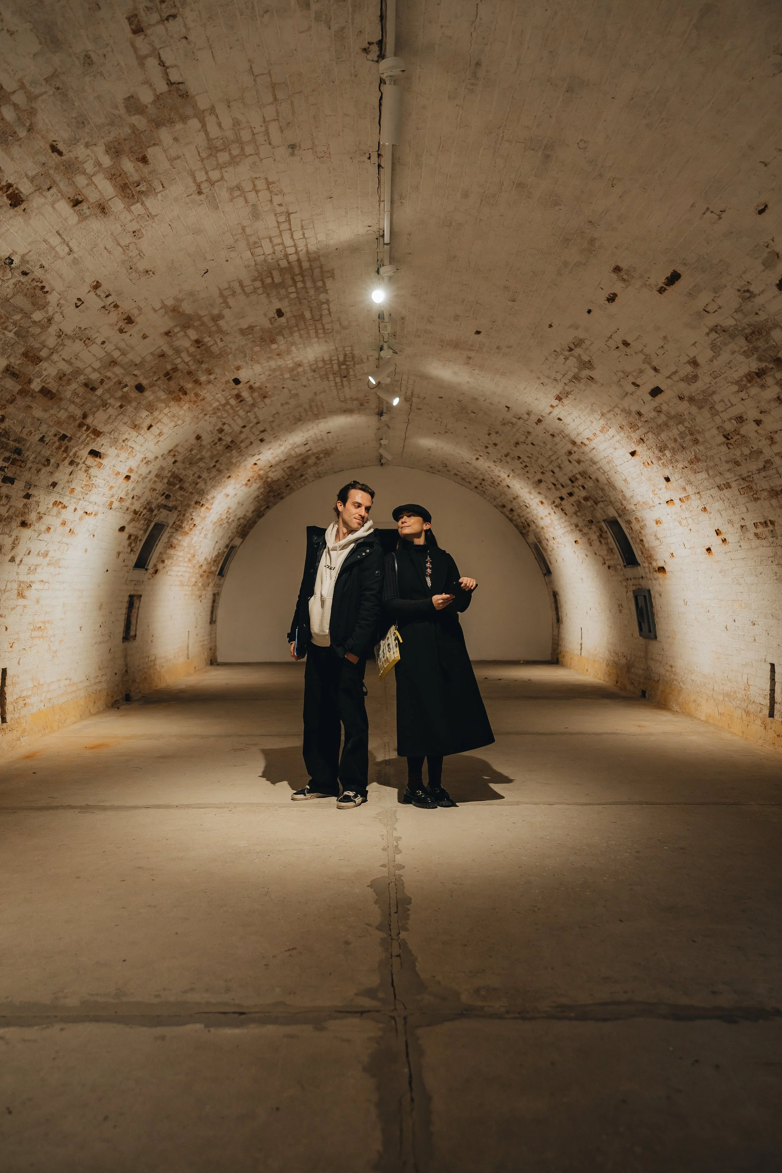 Two people standing inside a brick tunnel or archway, with warm lighting on the ceiling and floor.
