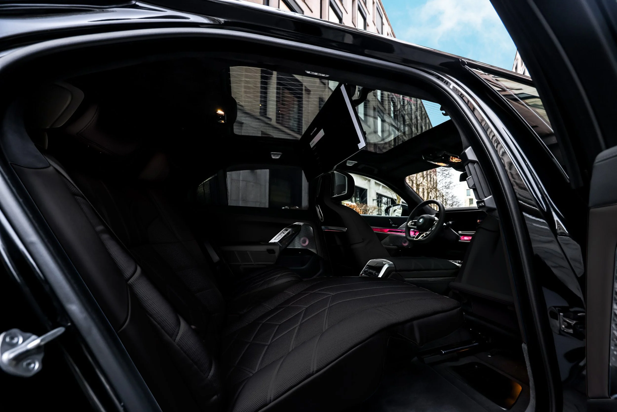 Interior of a modern, luxury vehicle with black seats, digital dashboard, and a large screen mounted on the ceiling. The vehicle is parked on a city street with buildings and a blue sky visible outside.