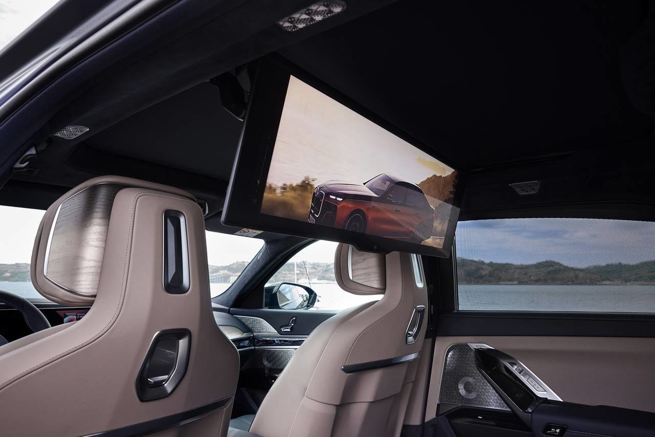 Interior of a luxury car showing beige leather seats, a dashboard with control panel, and a fold-down rear entertainment screen displaying an image of a red SUV driving through a landscape with hills and a cloudy sky.