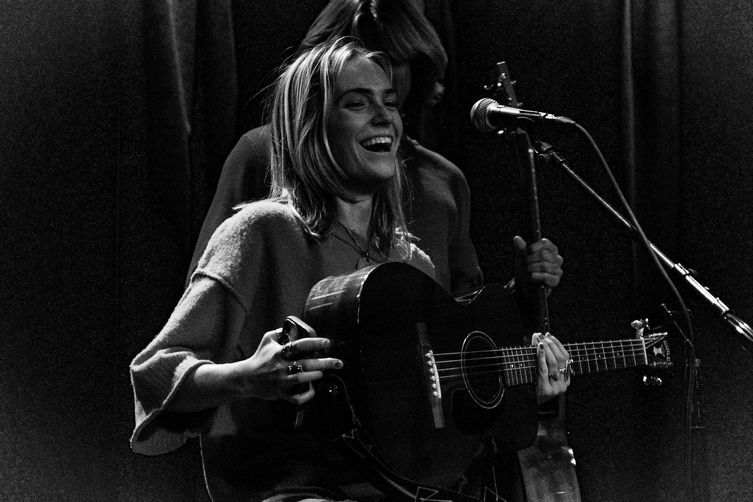 Maggie Nichols playing the guitar and singing into a microphone on stage, smiling and enjoying the performance, in black and white at SoHo House Santa Barbara.