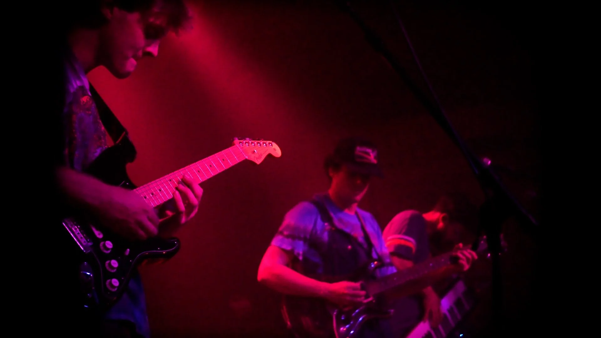Three musicians playing guitars on stage under pink and purple lights.