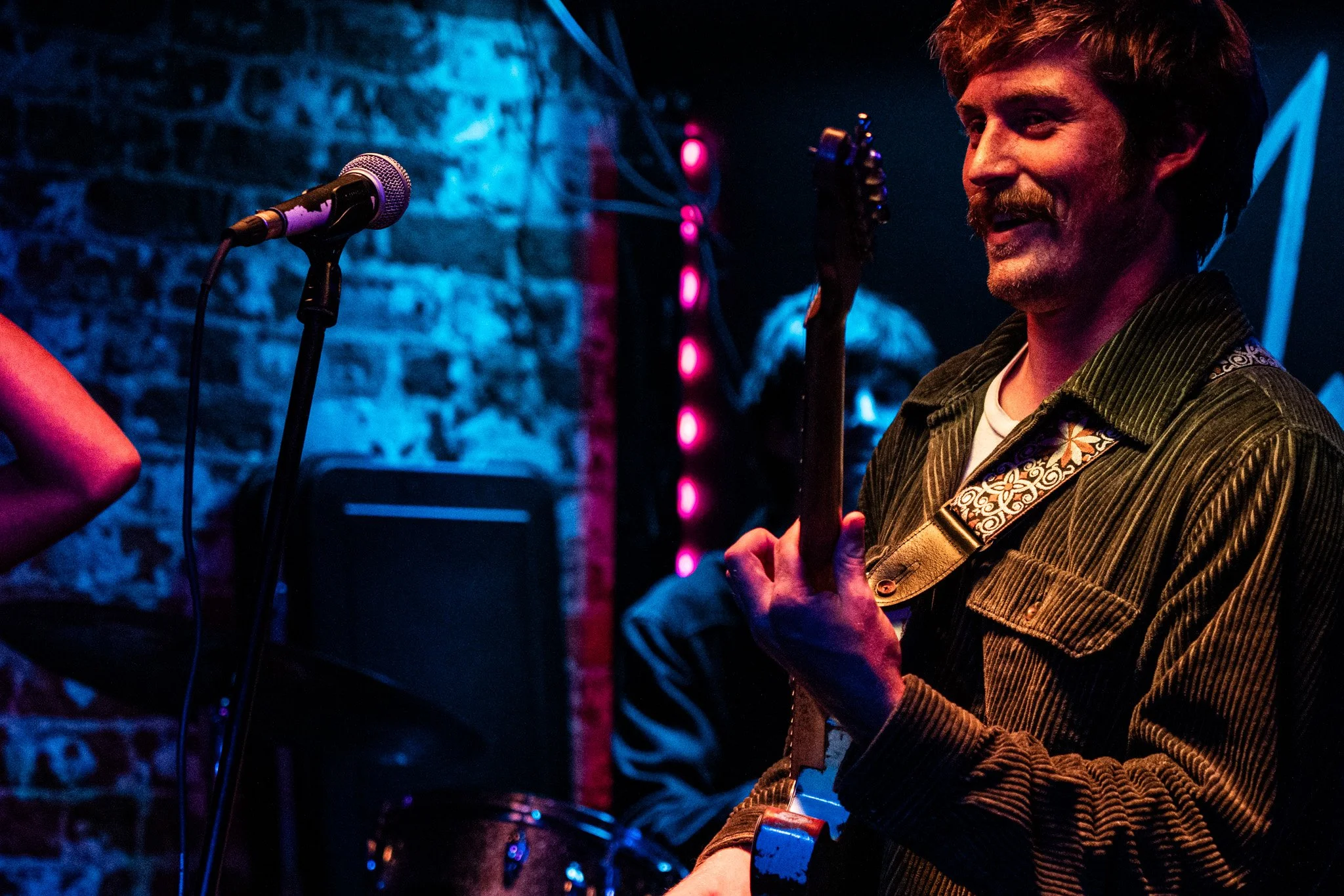 A man with a beard and curly hair playing an electric guitar on stage, with a microphone and a person playing drums in the background, under blue and pink stage lights.