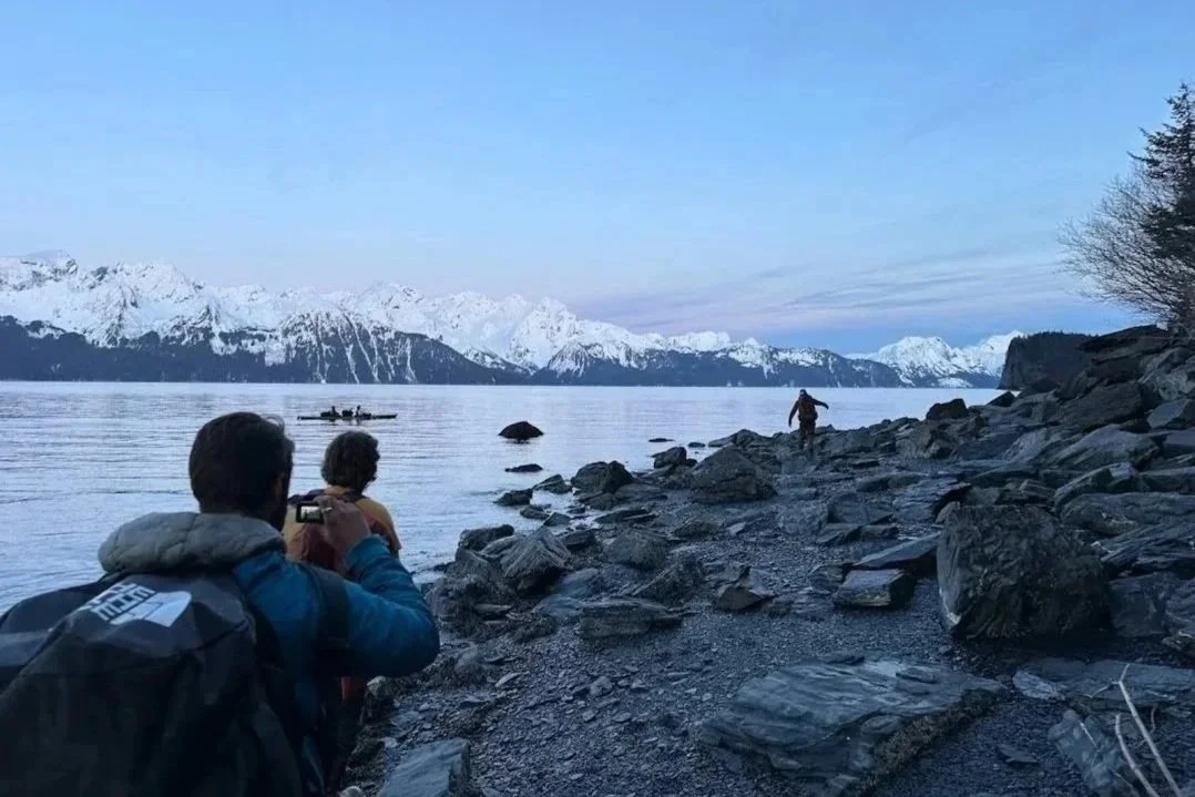 People on rocky shoreline taking photos of snow-capped mountains and a lake.