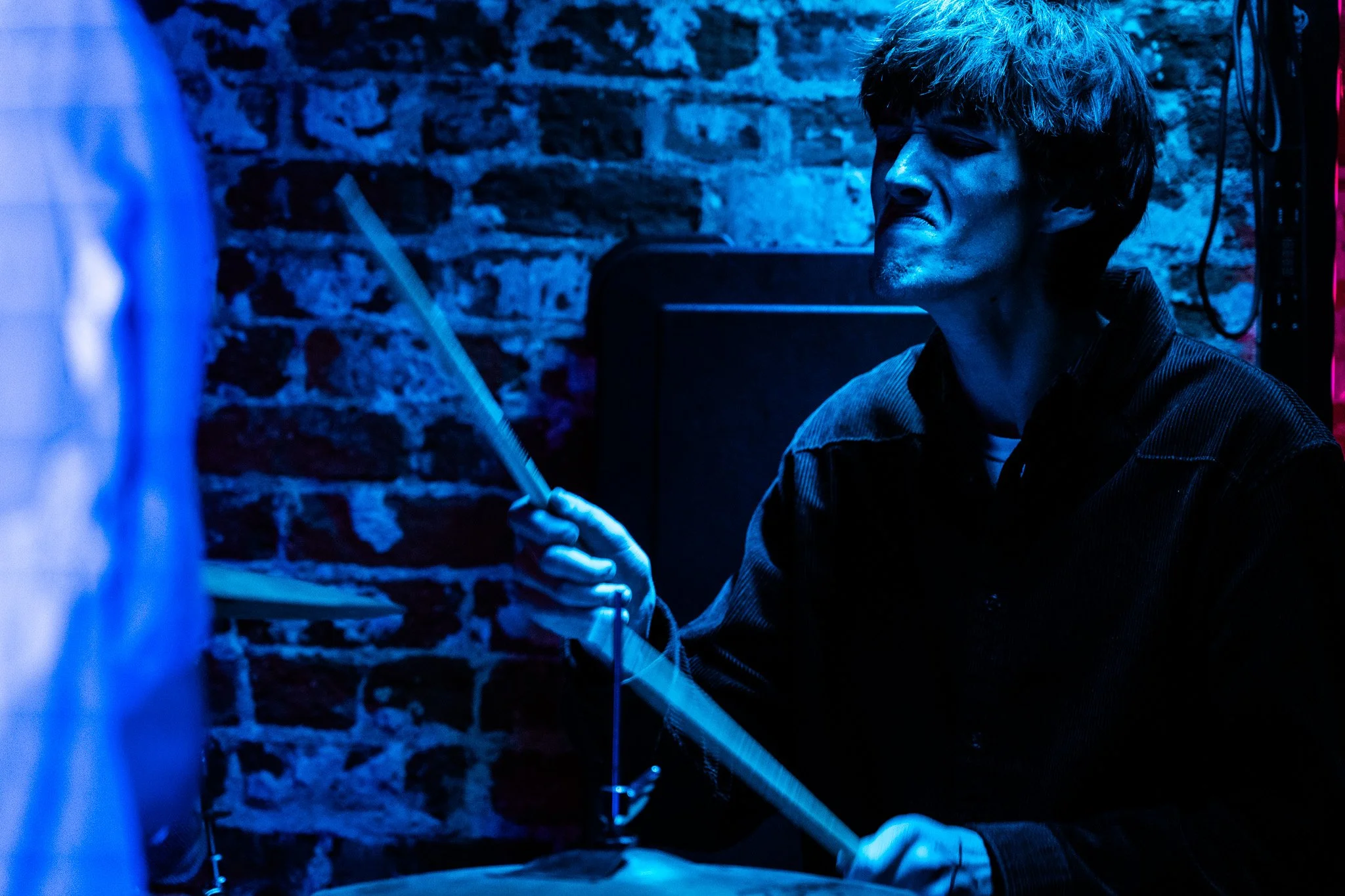 A man playing drums in a dark room illuminated by blue lighting, with brick walls in the background.