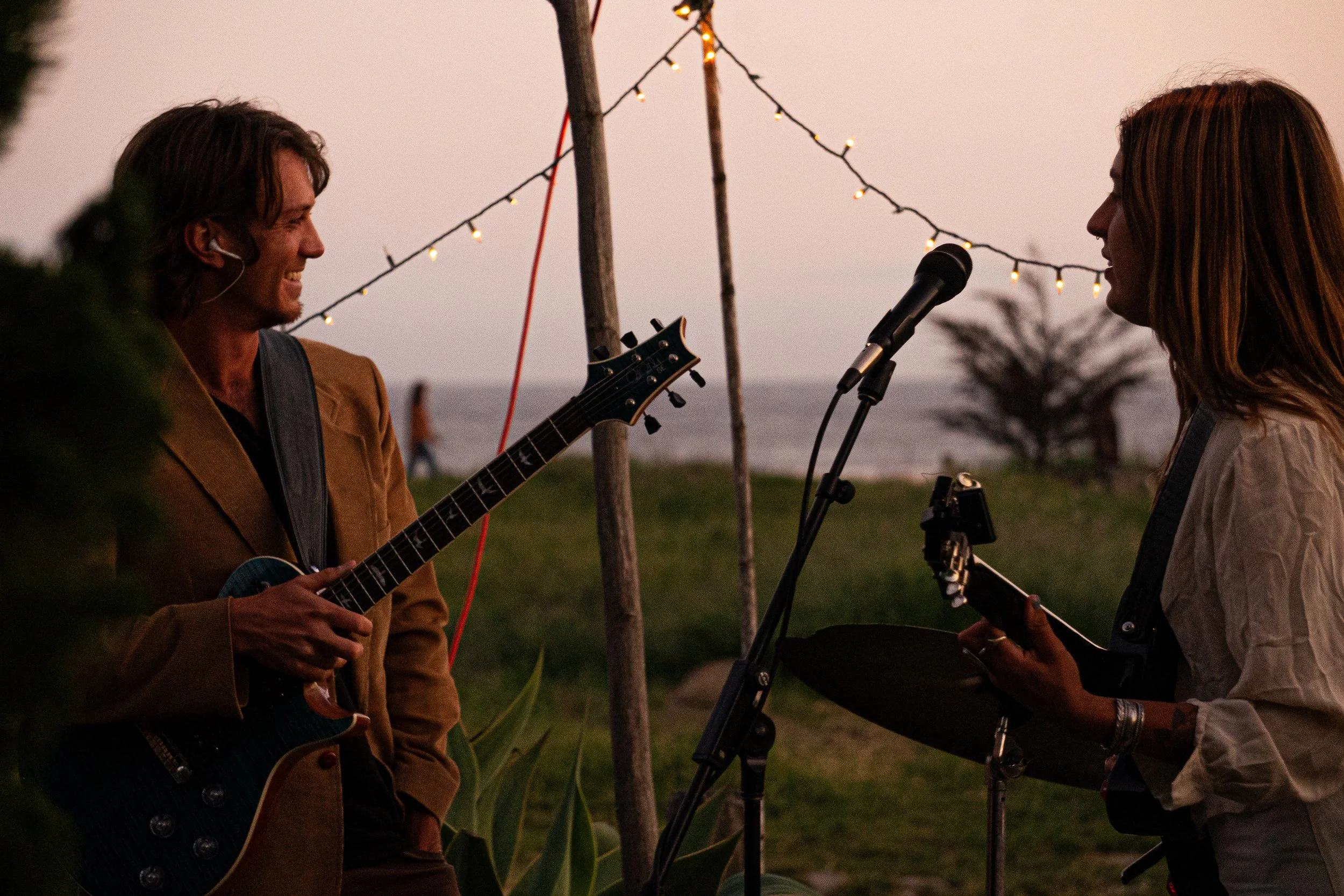 Two musicians perform outdoors at sunset, one playing an electric guitar and the other a percussion instrument, with string lights hanging above them, near the coast with a tree and grassy area in the background.